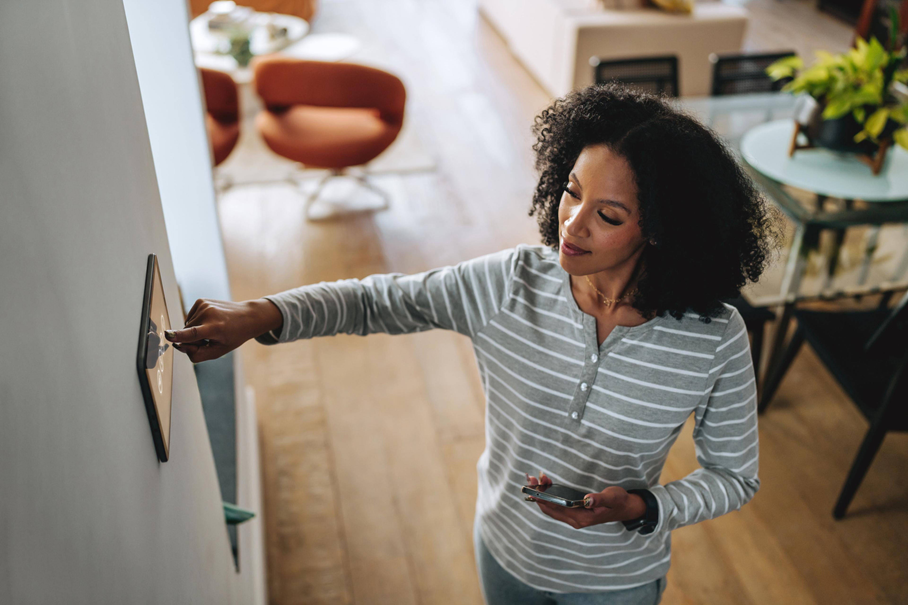 Eine Frau justiert in einem hellen, modernen Wohnzimmer einen an der Wand montierten intelligenten Thermostat, während sie ein Smartphone in der Hand hält.