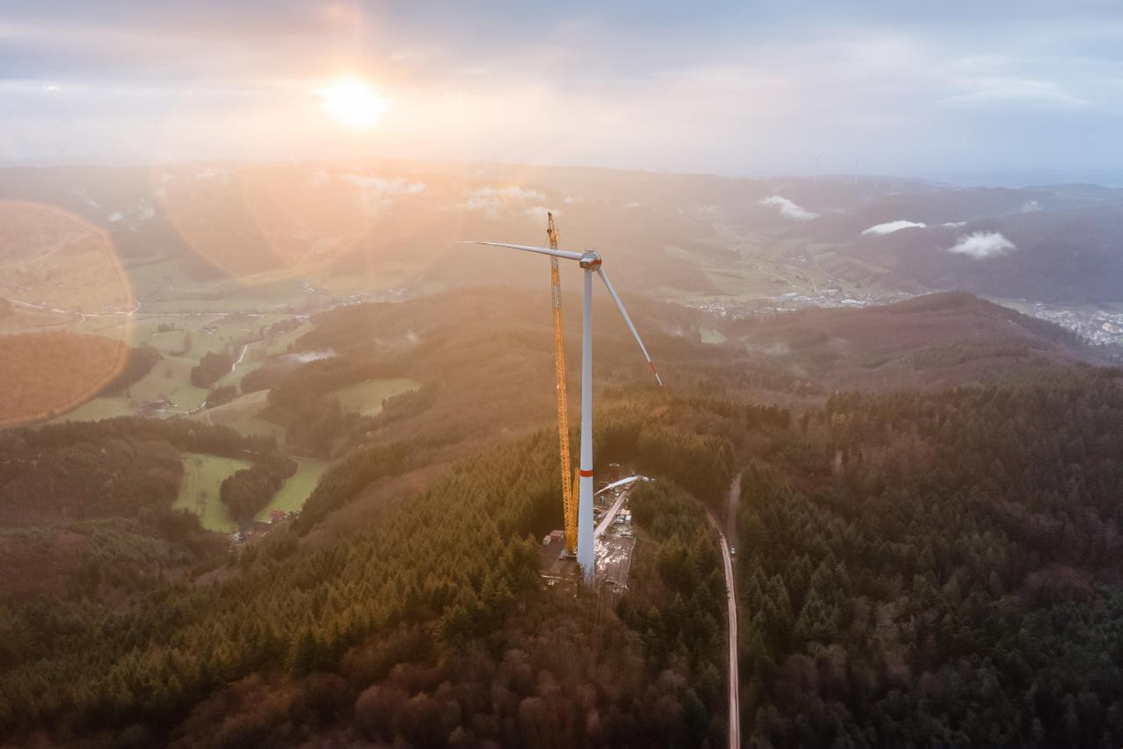 Aerial view of a wind turbine under construction on a wooded hilltop, with a crane nearby. Sun setting over hilly landscape in the background.