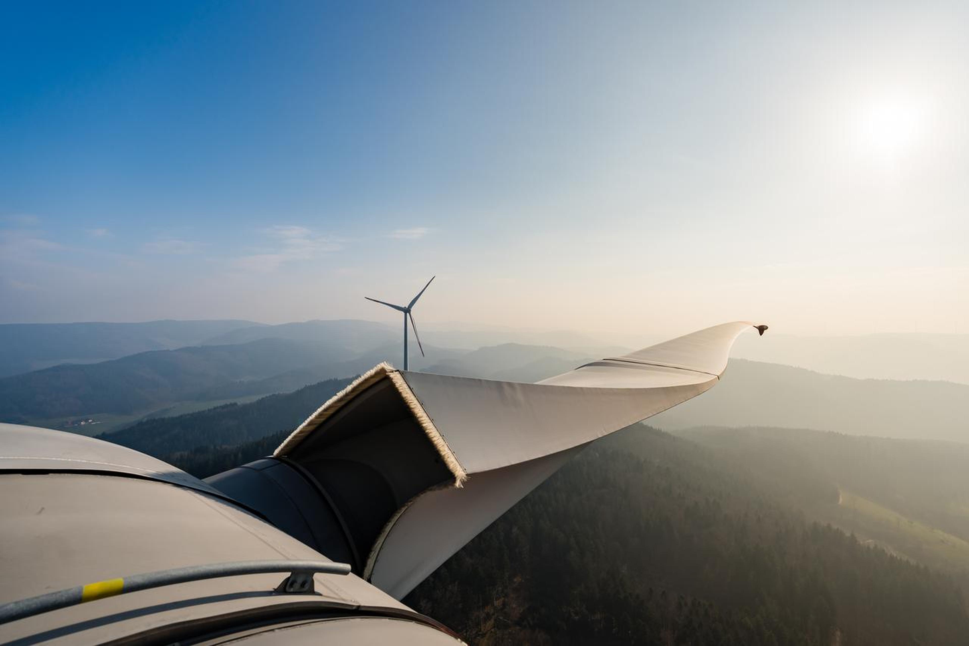 Close-up view of a wind turbine blade with misty mountains and another turbine in the background under a clear blue sky.