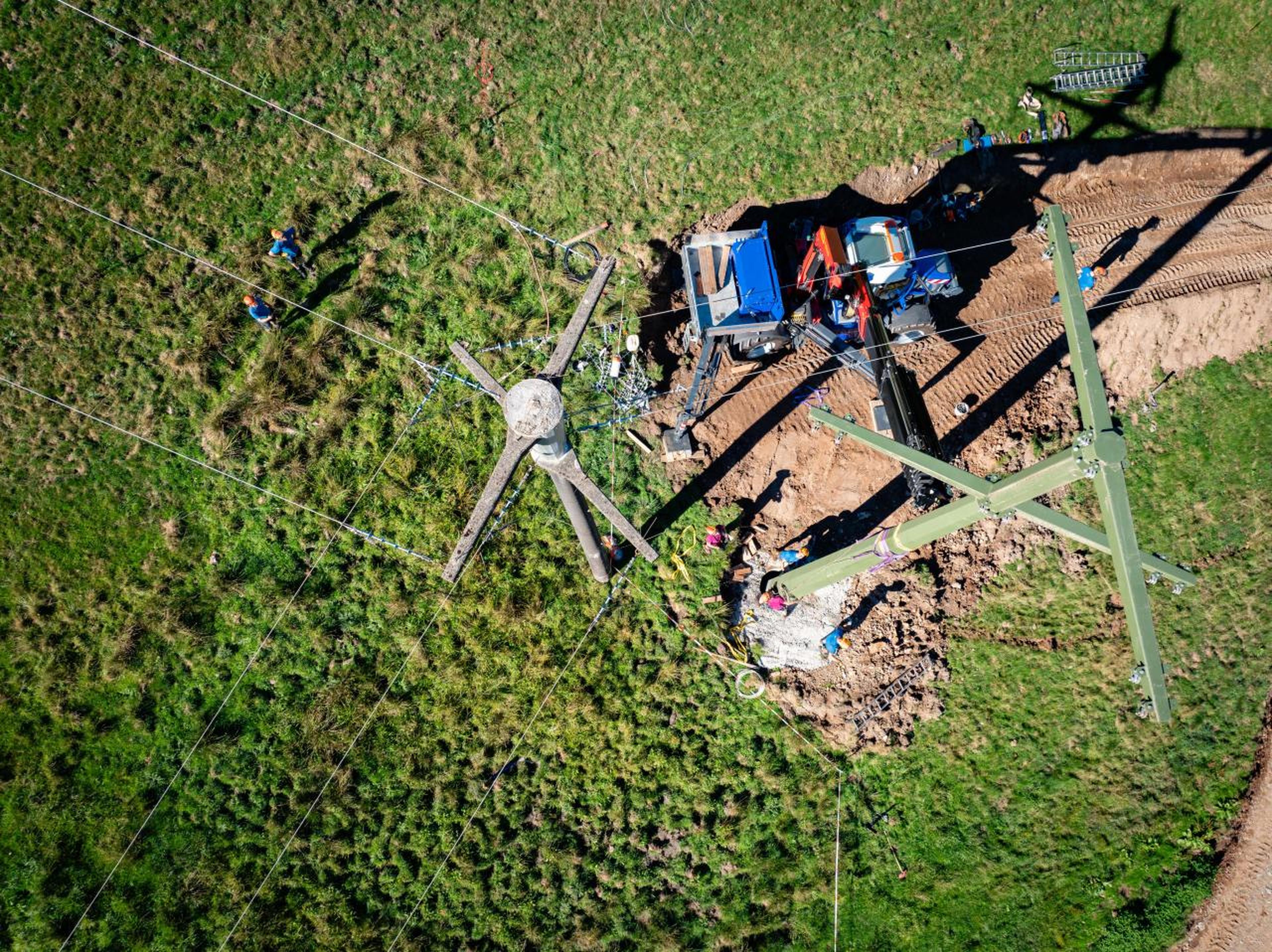 Aerial view of workers and machinery assembling a green power pylon in a grassy field with power lines radiating outward