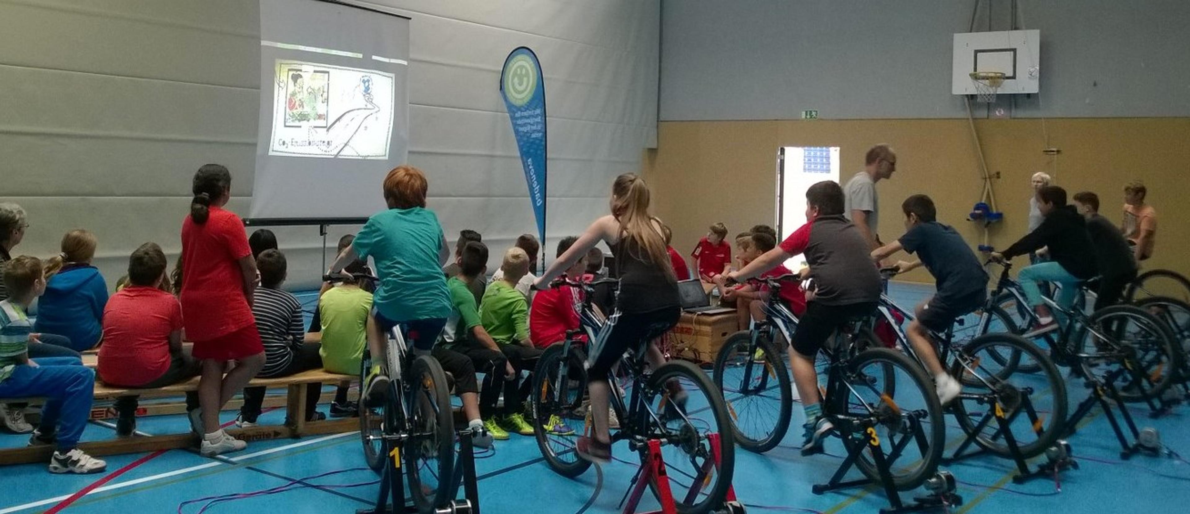 Kinder in einer Turnhalle treten auf stationären Fahrrädern in die Pedale, während sie eine Präsentation auf einem Bildschirm verfolgen, umgeben von ihren sitzenden Klassenkameraden.