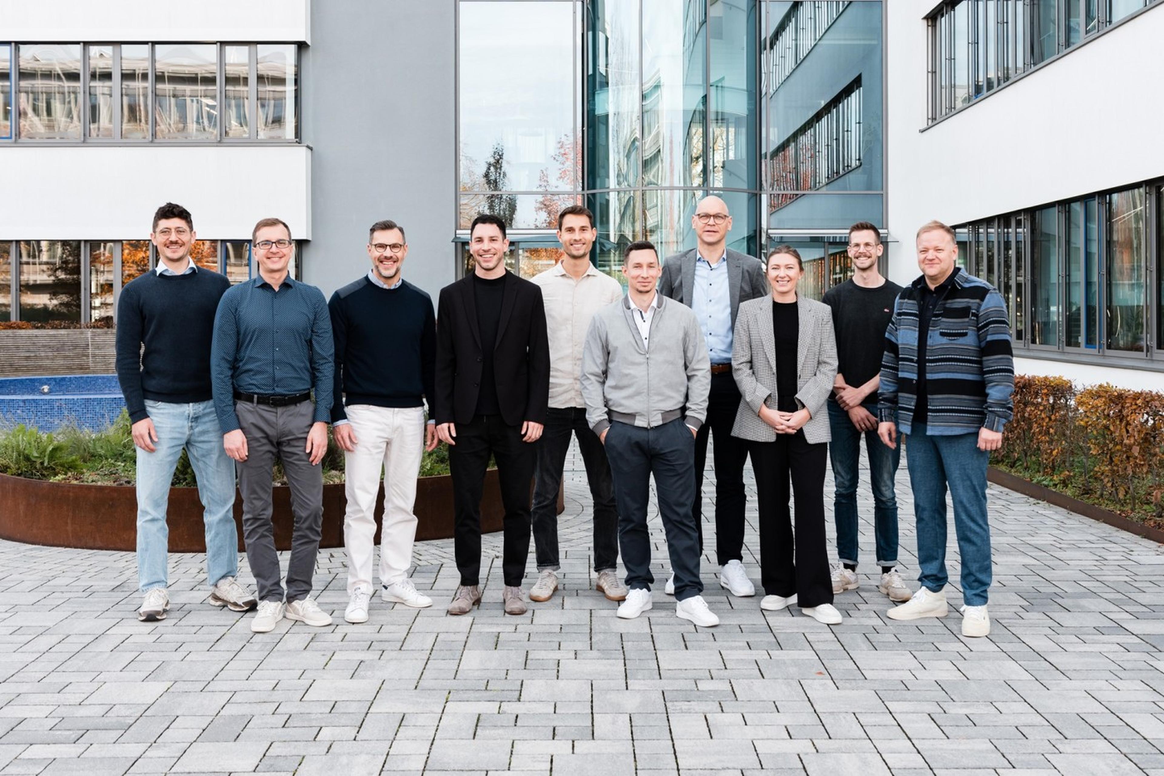 A group of eleven people standing outside a modern building, smiling. They are casually dressed, with a mix of men and women.