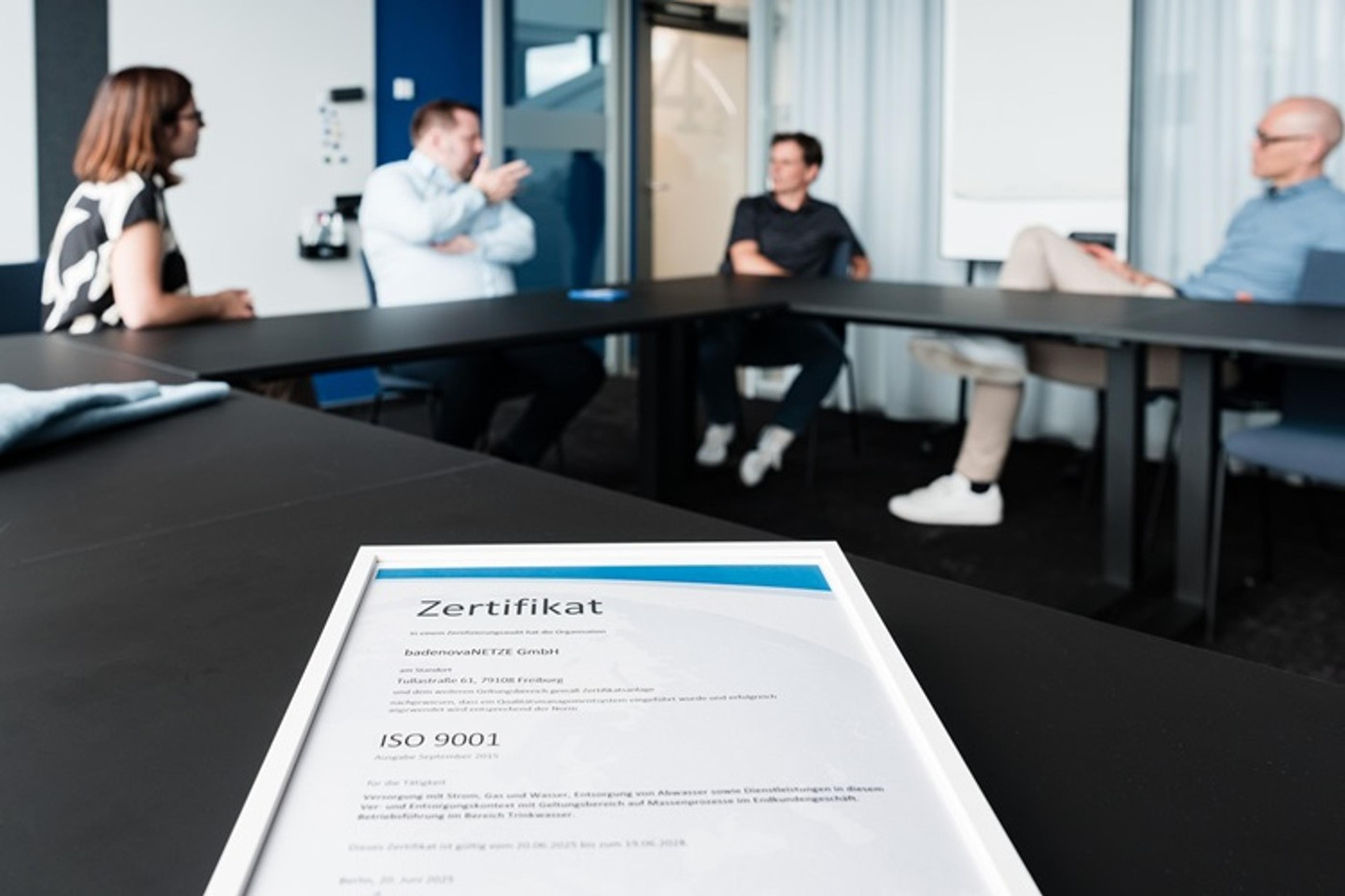 Eine Gruppe von vier Personen von BadneovaNETZE und Qualitätsmanagementbeauftragte, sitzt um einen Konferenztisch herum und diskutiert. Im Vordergrund ist ein Zertifikat mit der Aufschrift „ISO 9001” zu sehen.