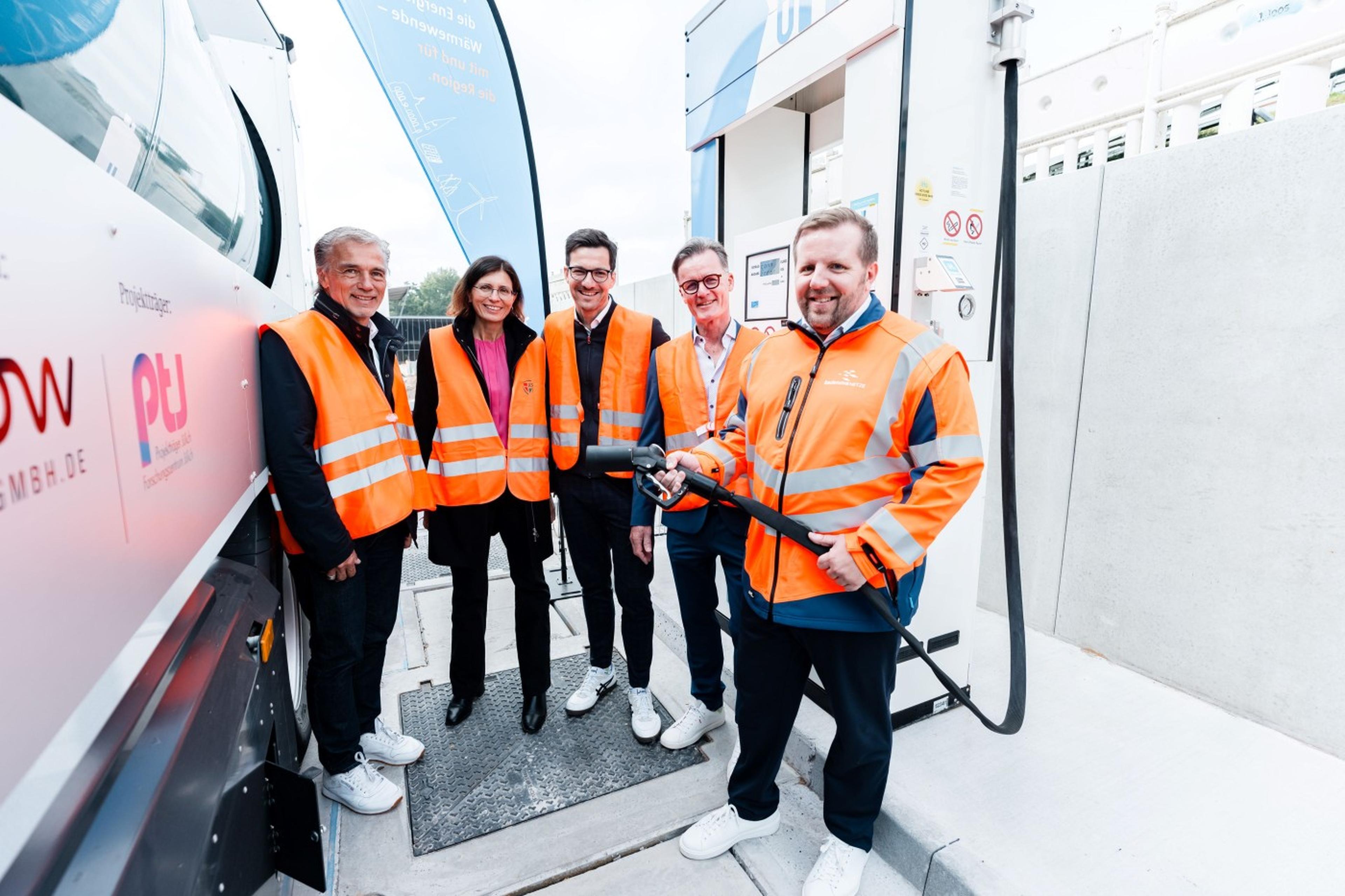 Fünf Personen stehen vor einer Wasserstoff-Tankstelle und lächeln in die Kamera. Eine Person hält einen Zapfhahn, als ob sie ein Fahrzeug betanken würde. Im Hintergrund sind ein Banner und Teile der Tankstelle sichtbar.