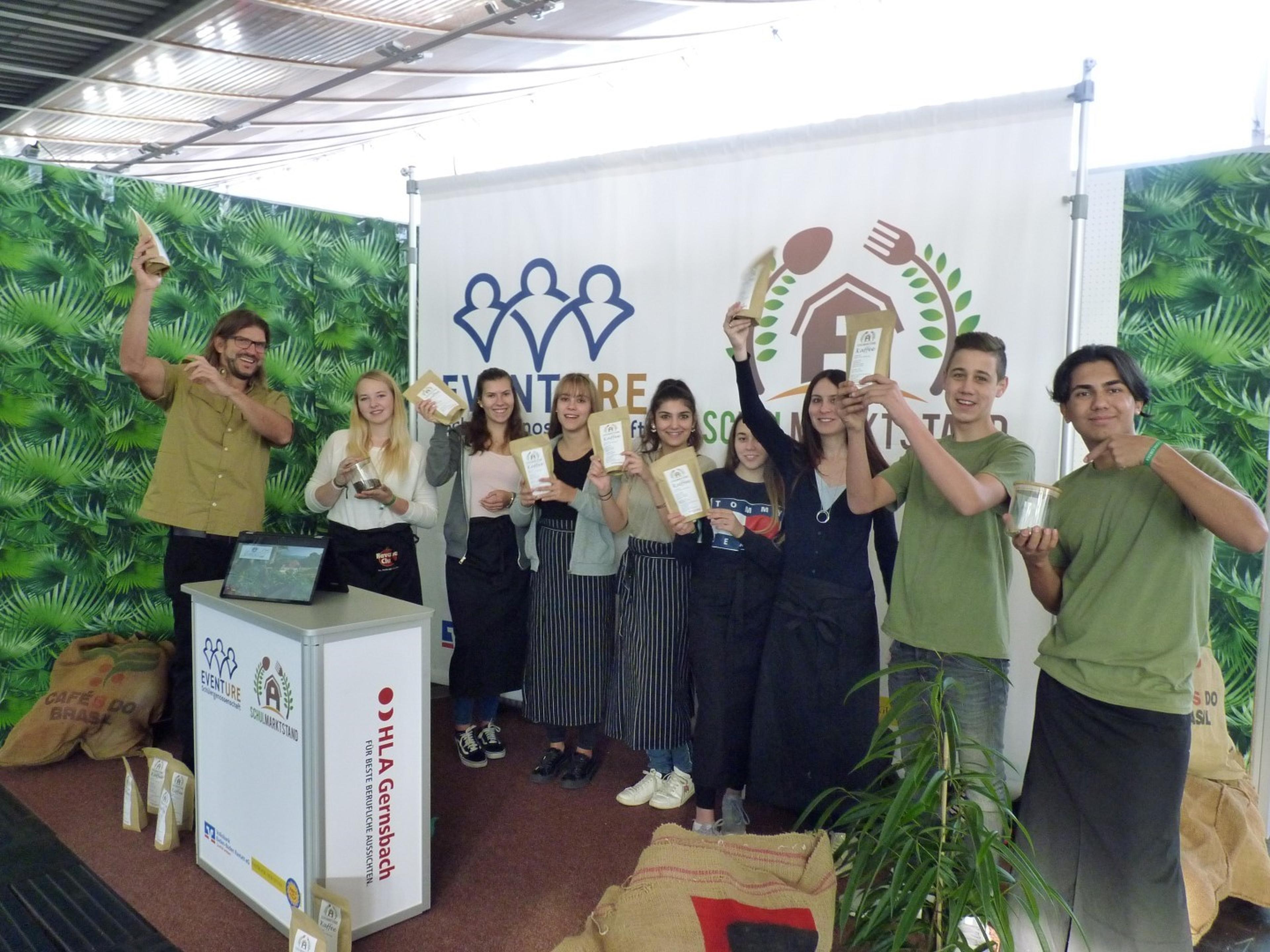 Eine Gruppe von Menschen, die lächeln und Kaffeetüten in den Händen halten, vor einem Stand mit tropischem Hintergrund und einem Logo.