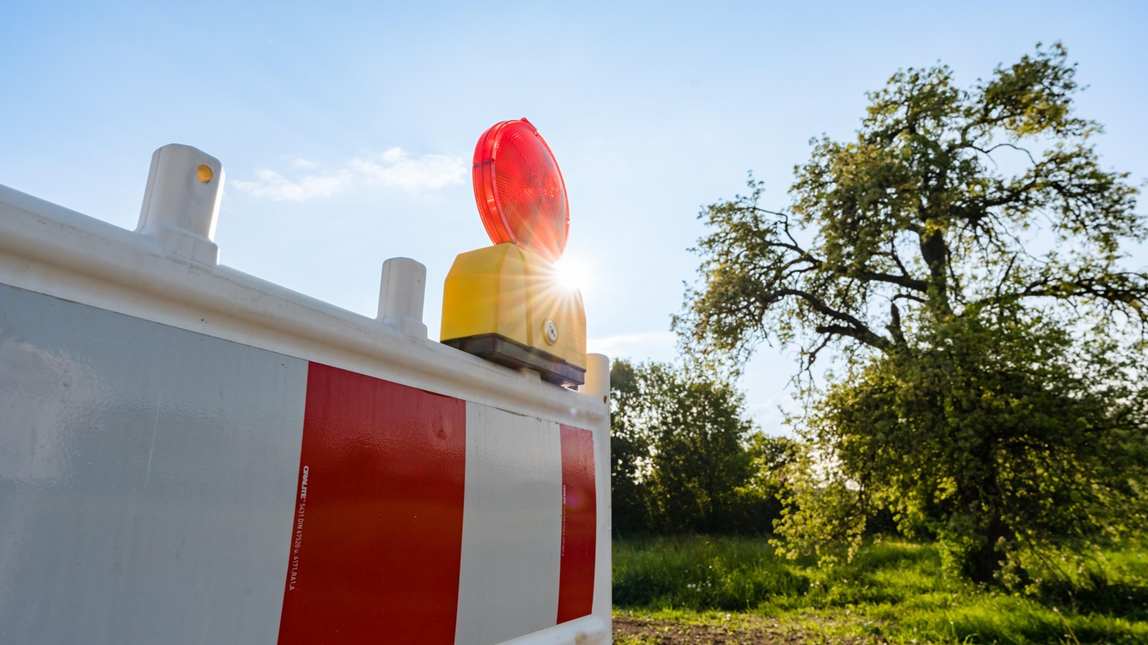 Road barrier with red and white stripes and a red warning light, set against a sunny, tree-lined landscape.