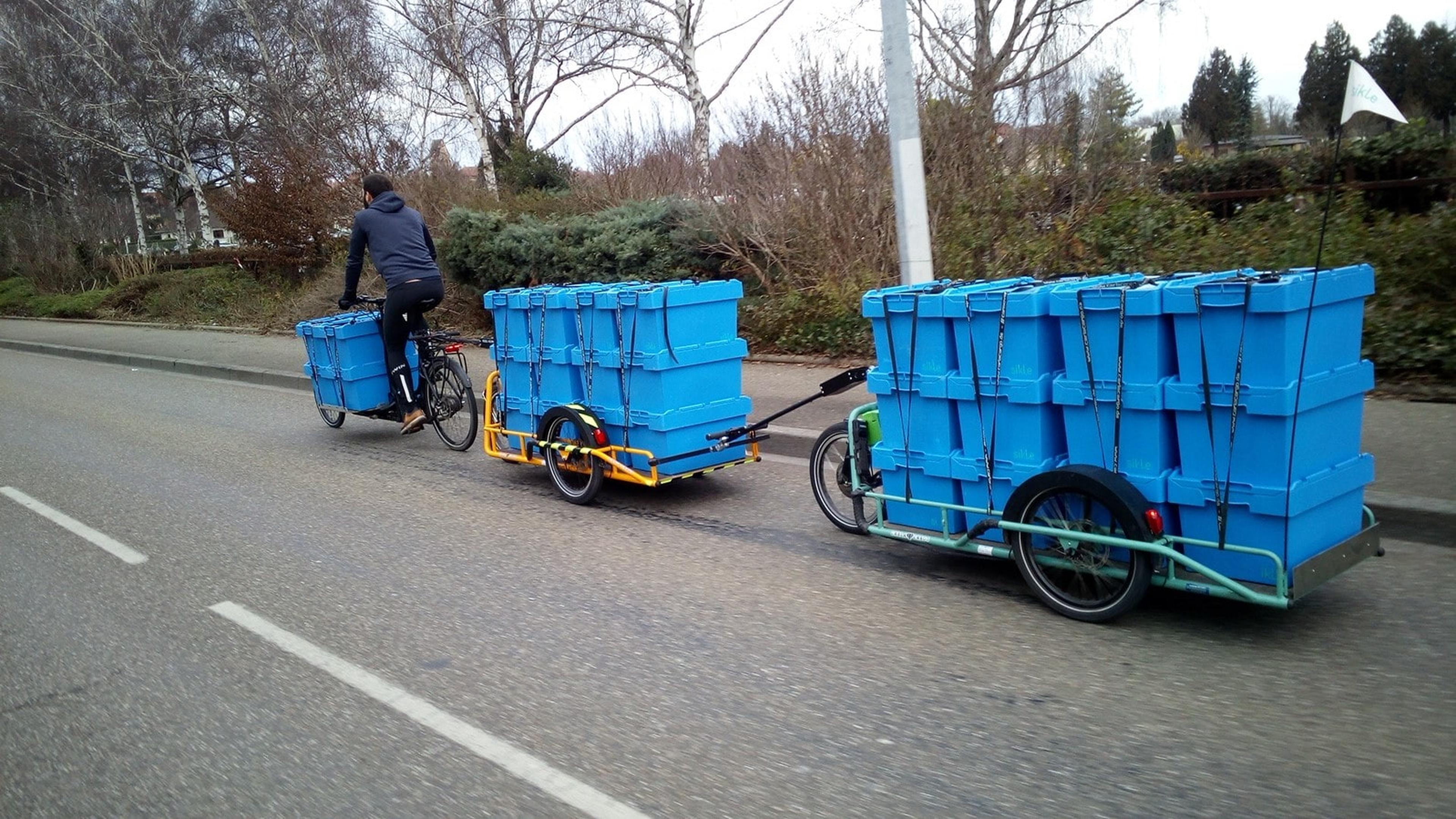 Person, die auf einer Straße Fahrrad fährt und zwei Anhänger mit gestapelten blauen Containern zieht, umgeben von Bäumen und Sträuchern.