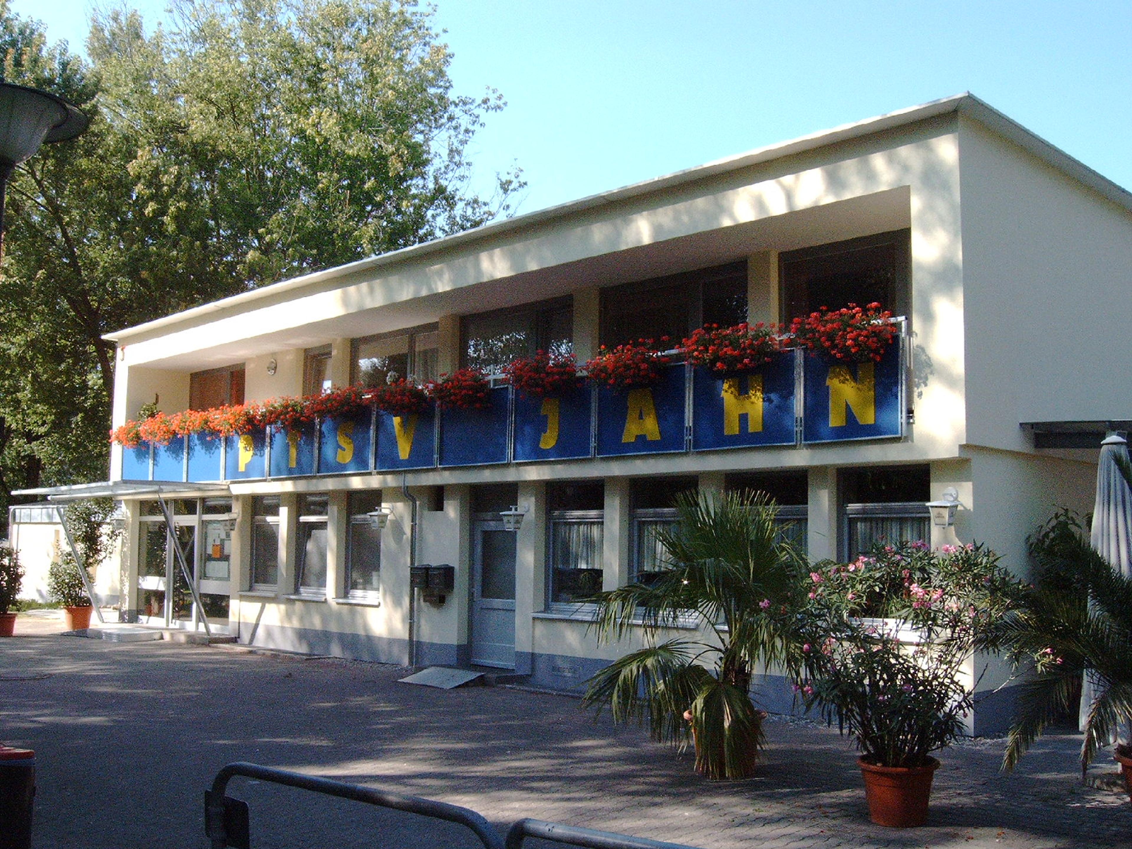 Ein zweistöckiges Gebäude mit der Aufschrift „PSV JAHN“, Blumenkästen und Topfpflanzen davor, vor dem Hintergrund von Bäumen und einem klaren Himmel.