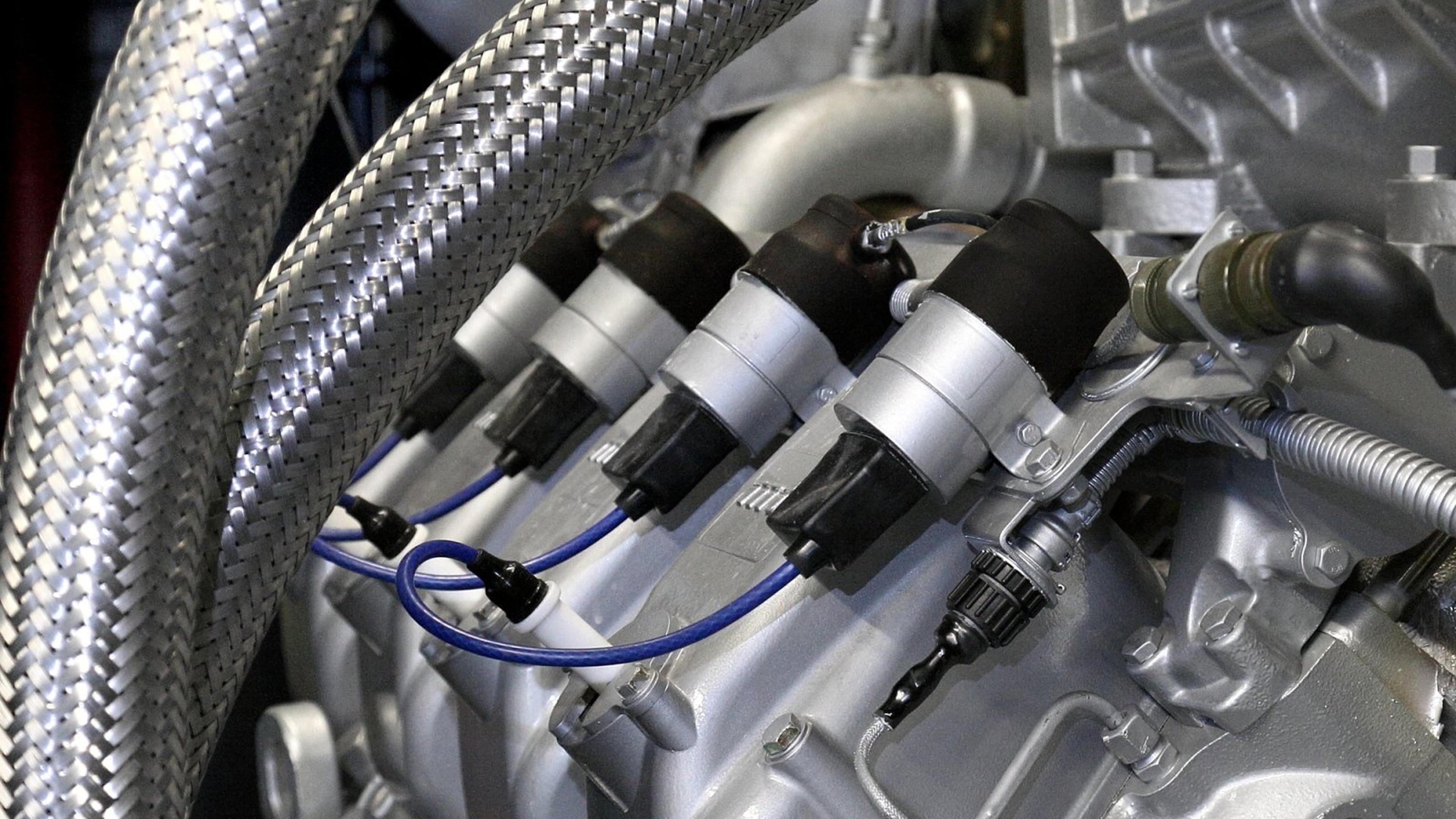Nahaufnahme eines Motors mit vier Zündkerzen, die durch blaue Kabel verbunden sind, umgeben von metallischen Gewebeschläuchen und mechanischen Bauteilen.