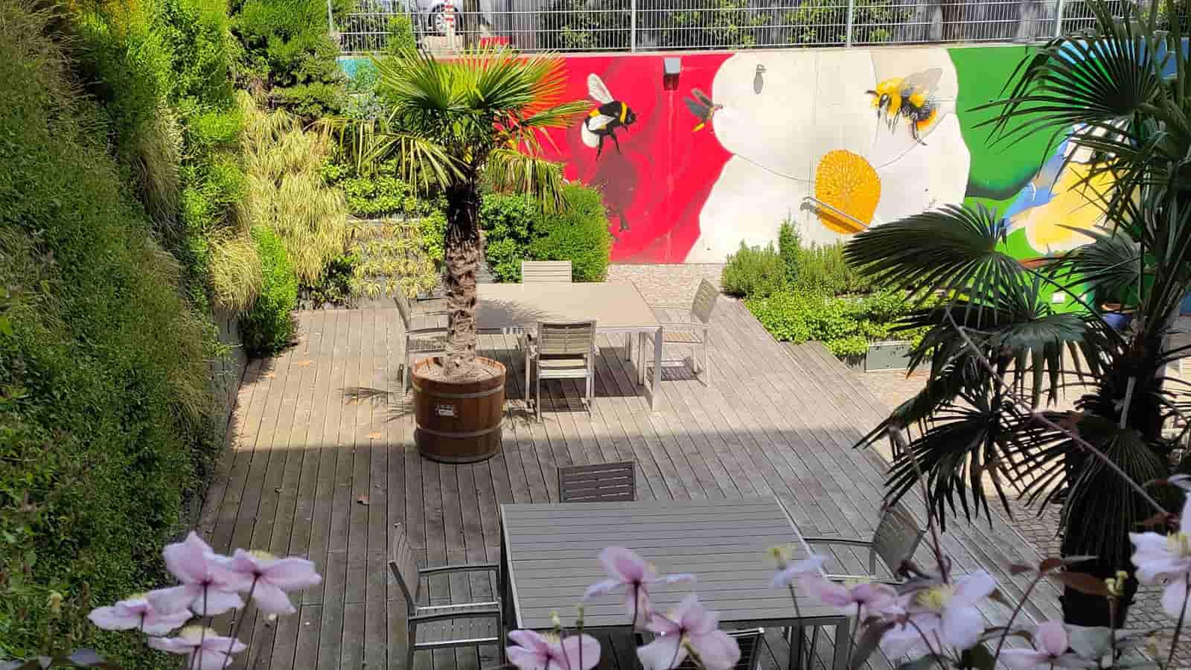 Außenbereich mit Holzterrasse, Tischen und Stühlen. Lebhafte Wandmalerei mit Blumenmotiven, umgeben von üppigem Grün und Palmen.