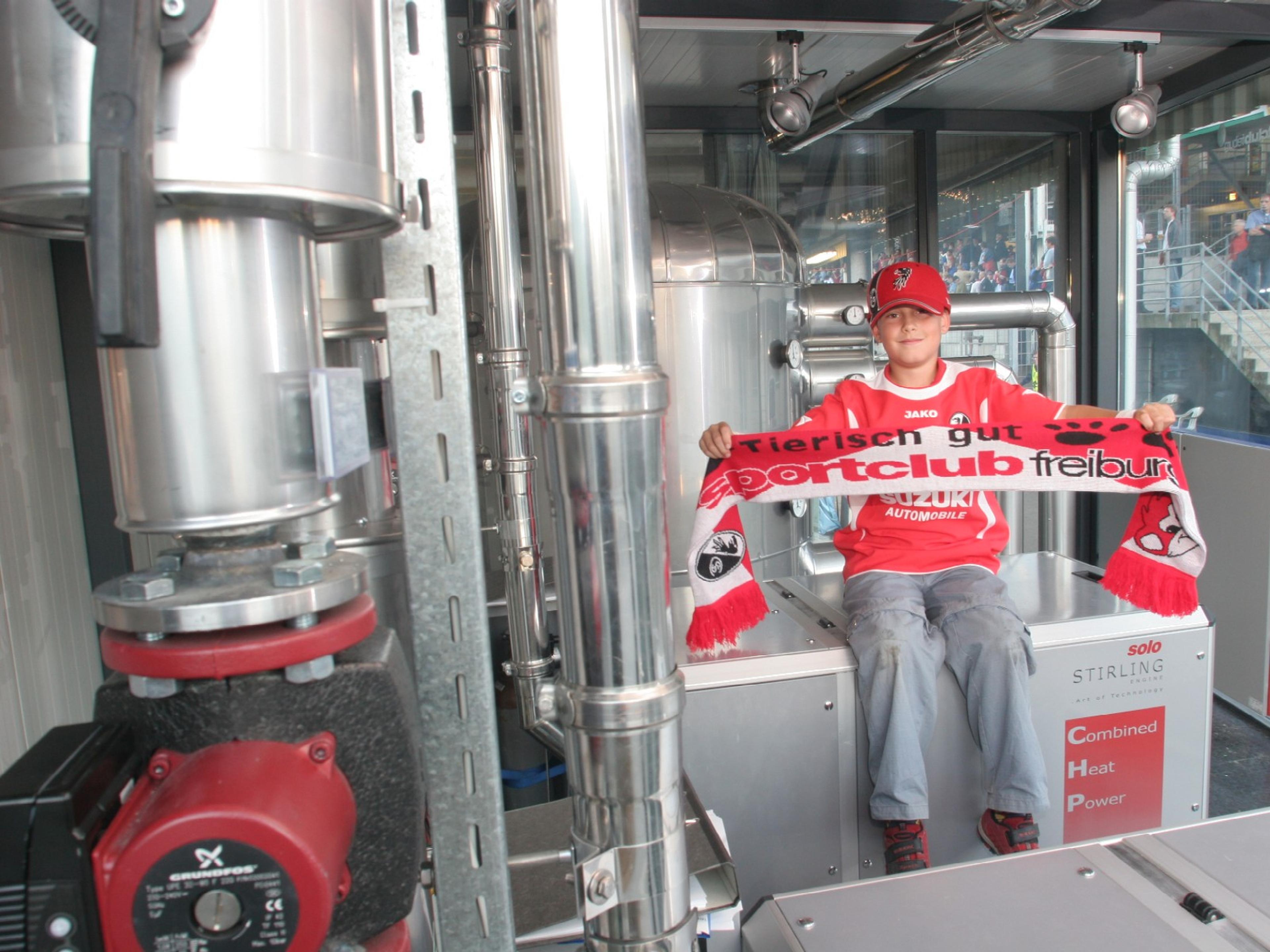 Young fan wearing a red cap and shirt holds a Sportclub Freiburg scarf, sitting on machinery in a technical setting.
