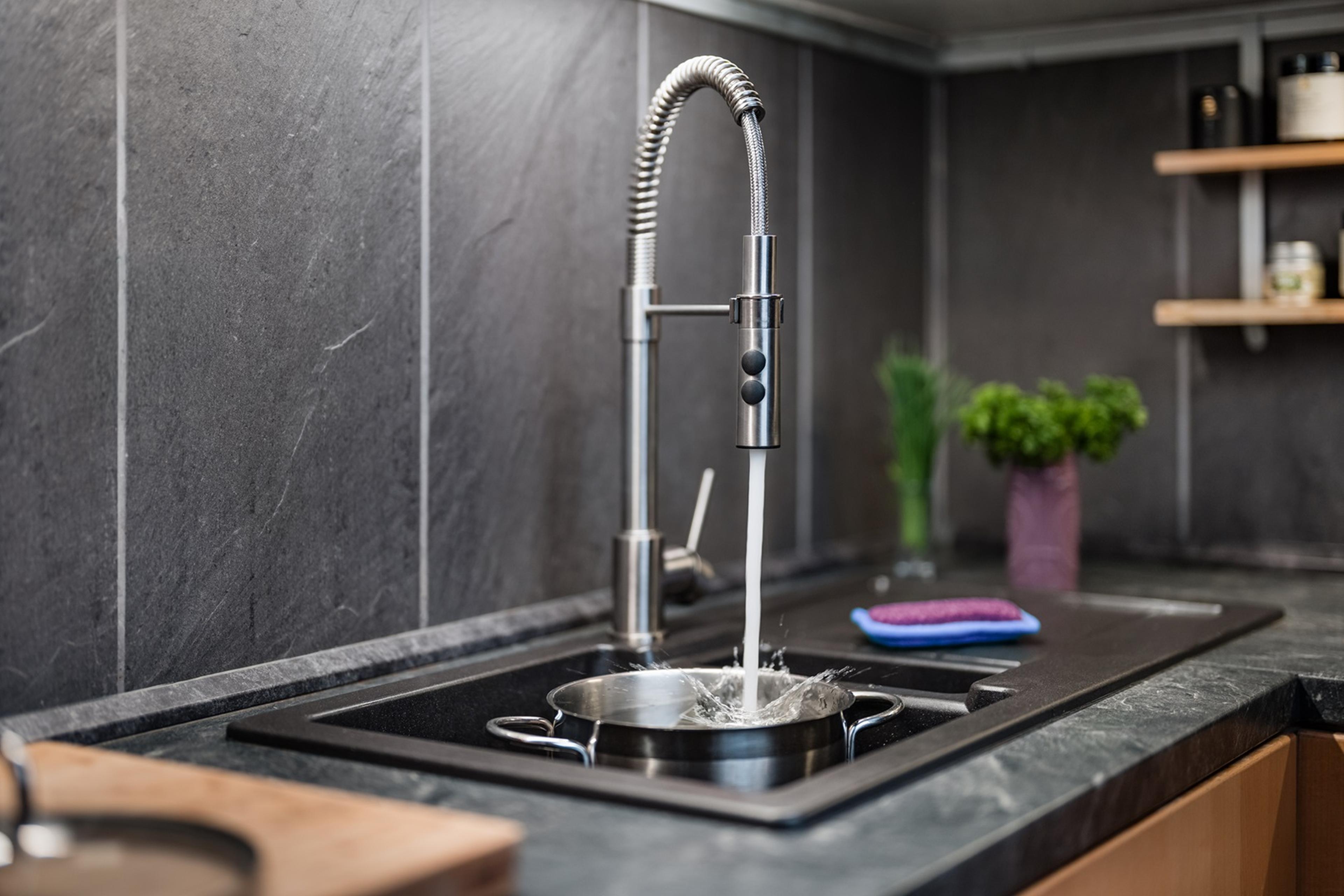 Water flowing from a modern, coiled kitchen faucet into a pot on a black countertop, with shelves and greenery in the background.