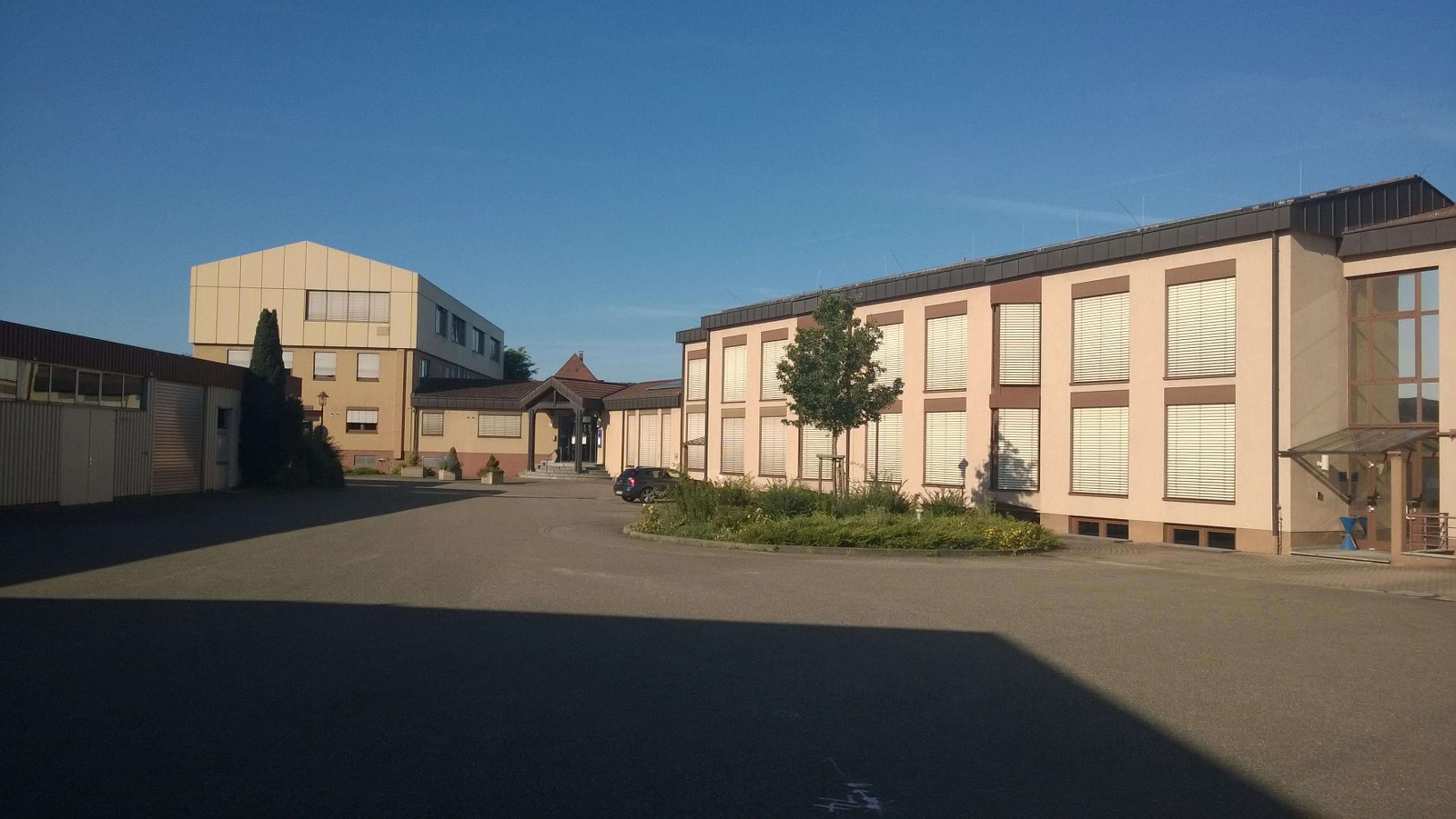 Industriekomplex mit mehreren beigen Gebäuden, großen Fenstern und einem kleinen Baum in der Mitte, unter einem klaren blauen Himmel.