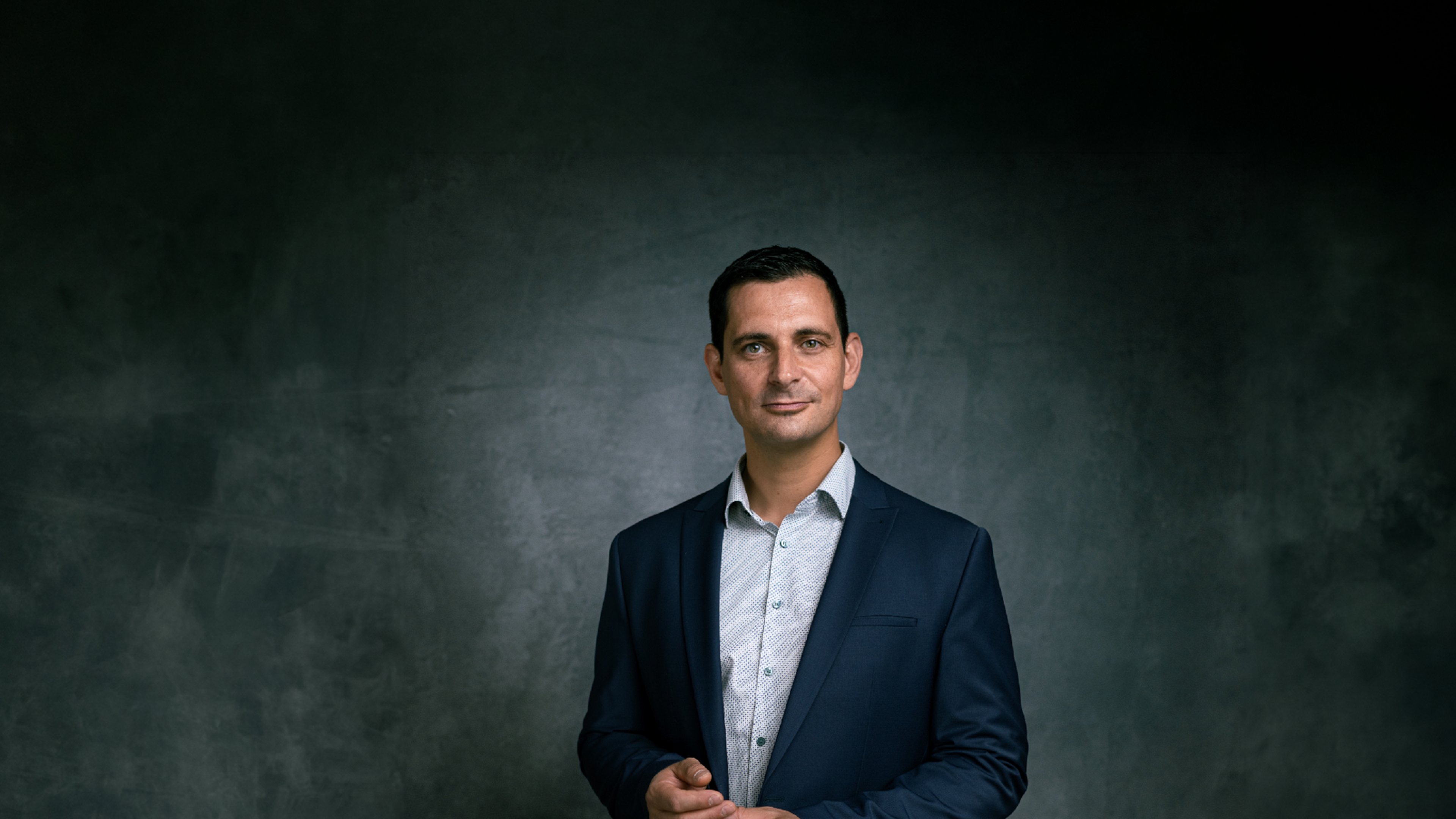 A man in a blue suit and light shirt stands against a dark textured background, posing with a neutral expression.