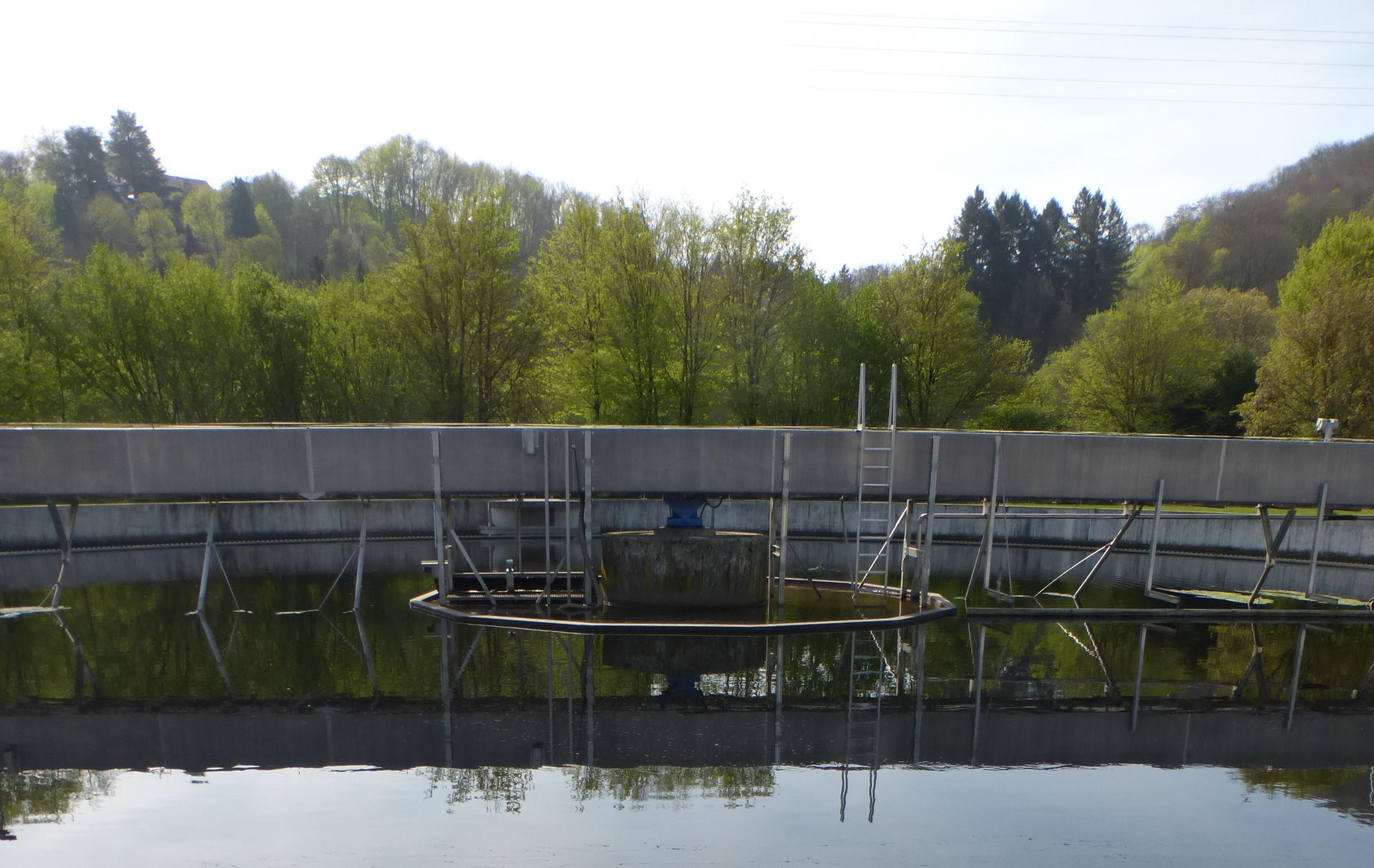 Eine kreisförmige Wasseraufbereitungsanlage mit Leitern und Geländern, umgeben von Bäumen und Hügeln unter einem klaren Himmel.