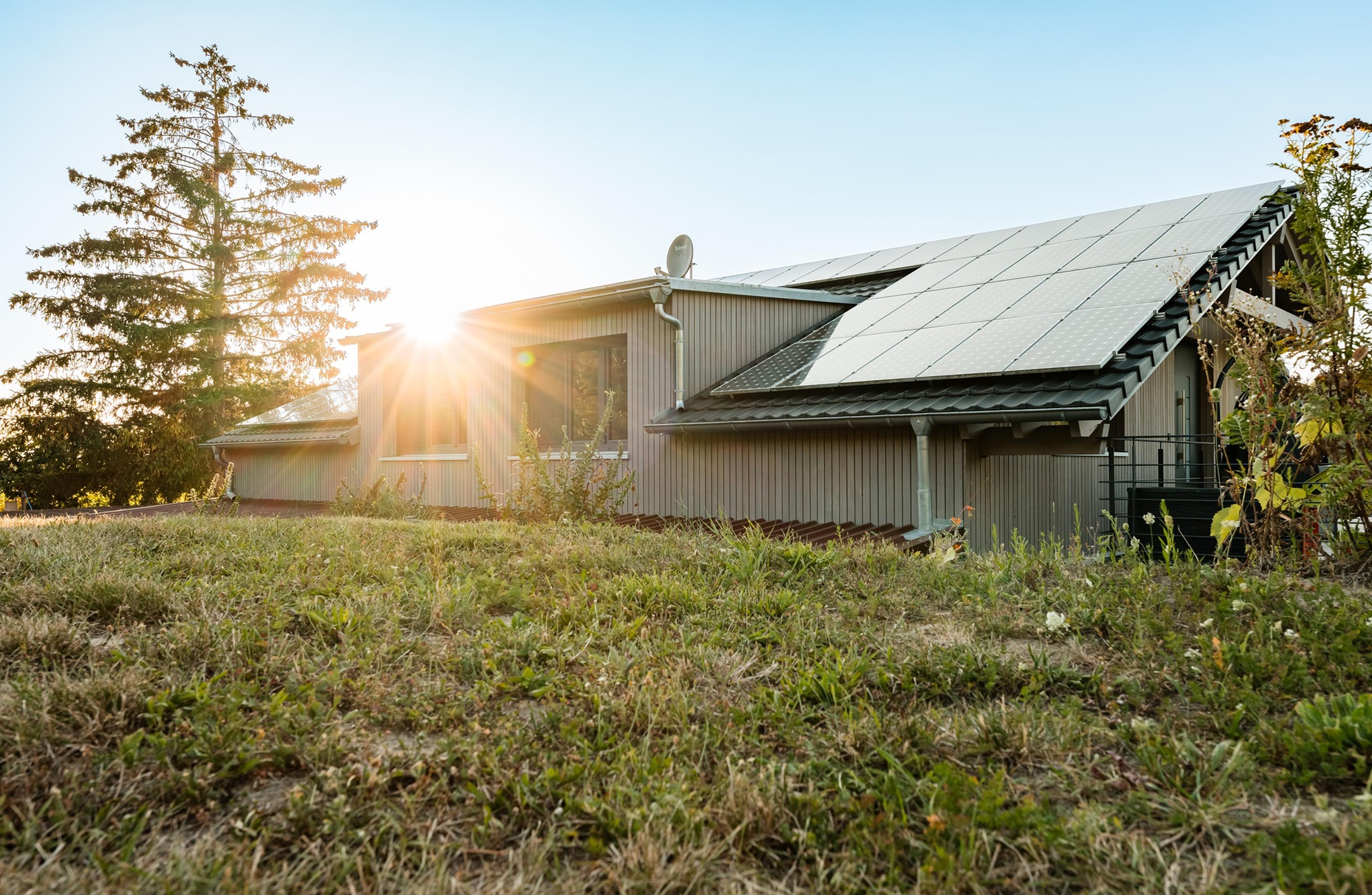 Ein graues Haus mit einem schwarzen Dach, das komplett mit Photovoltaik Paneelen bedeckt ist. Die Sonne schimmert auf das Dach, der Himmel ist blau. Im Vordergrund befindet sich eine Wiese.