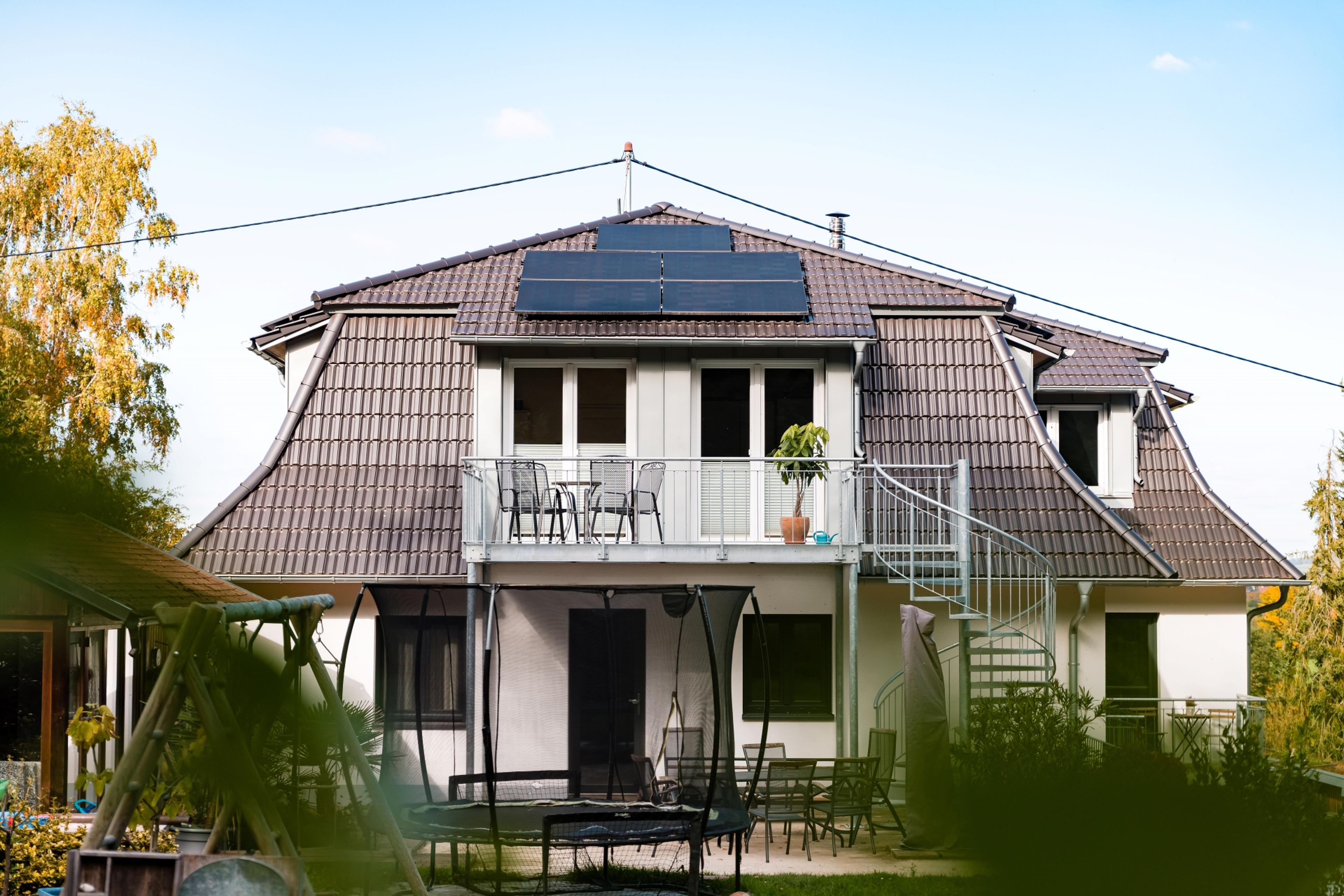 Ein weißes Haus mit einem grauen, tiefgezogenen Dack. In der Mitte des Dachs befinden sich mehrere Photovoltaik-Module. Darunter befindet sich ein Balkon mit einer Wendeltreppe in den Garten. Der Himmel scheint blau, die Bäume sind herbstlich gelb