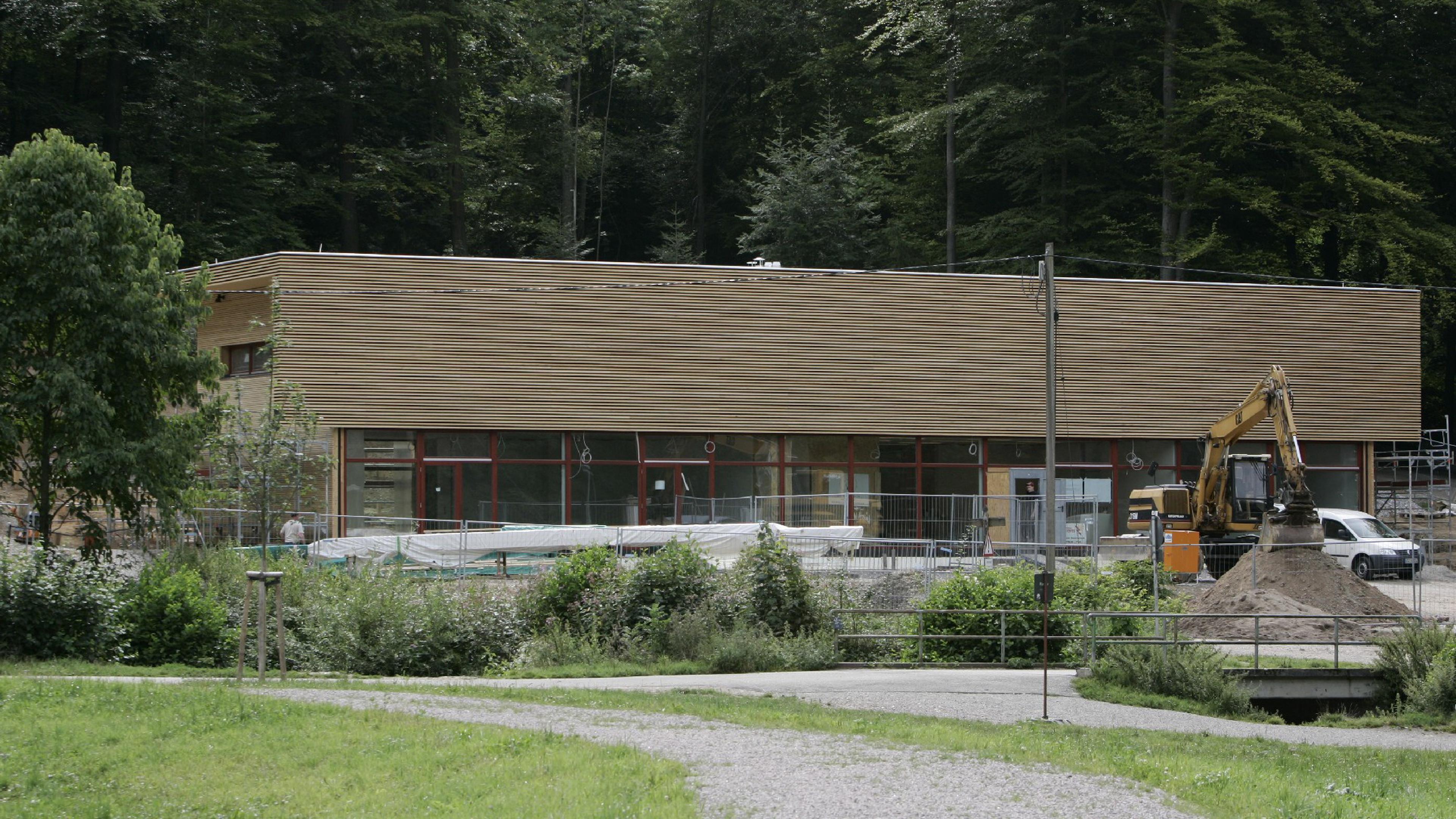 Ein modernes Holzgebäude, das gerade in einem Waldgebiet gebaut wird, mit einem Bagger und einem Erdhaufen im Vordergrund.