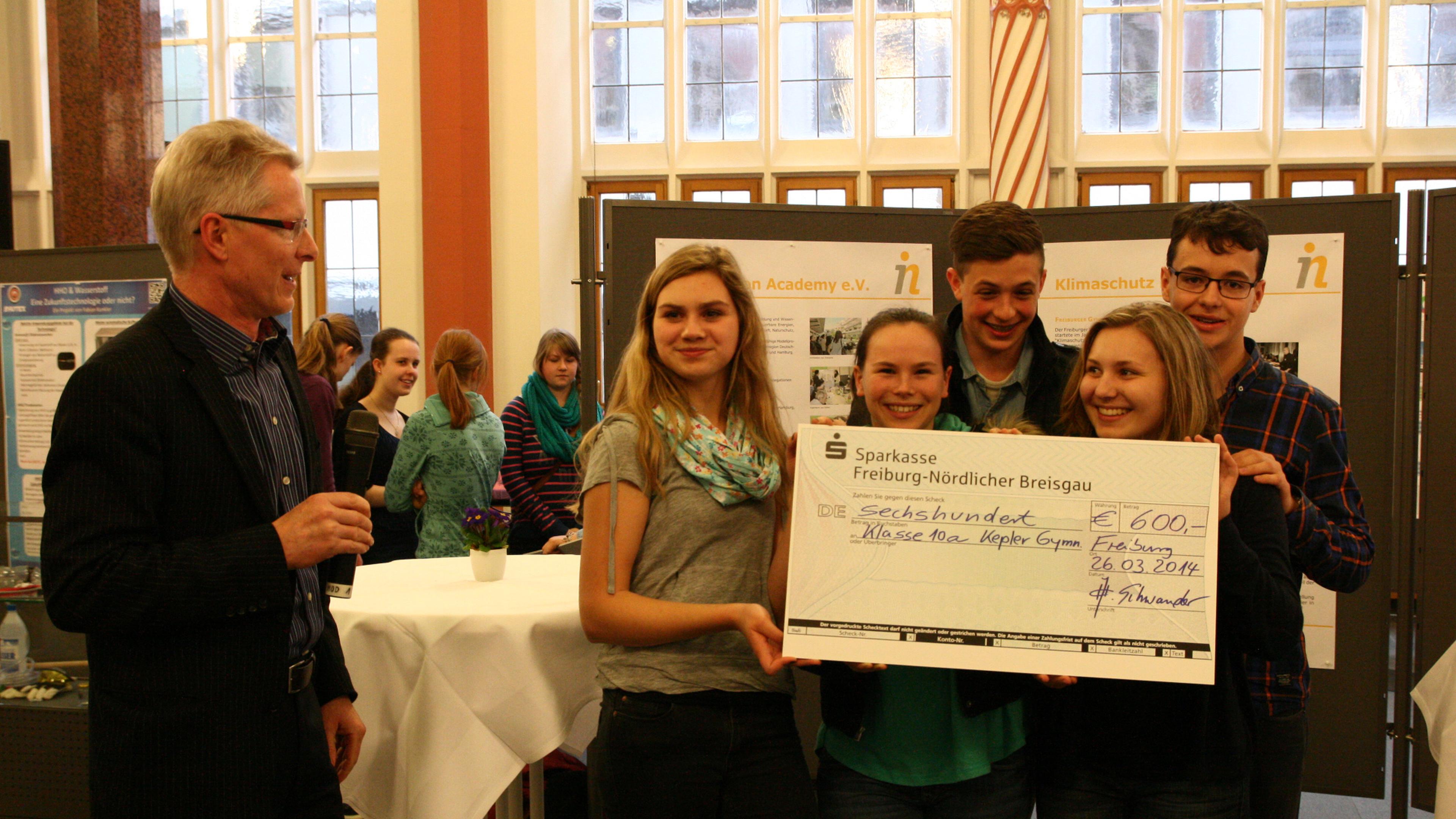 Eine Gruppe junger Leute, die bei einer Veranstaltung einen großen Scheck über 600 € in den Händen hält, während neben ihnen ein Mann ins Mikrofon spricht.