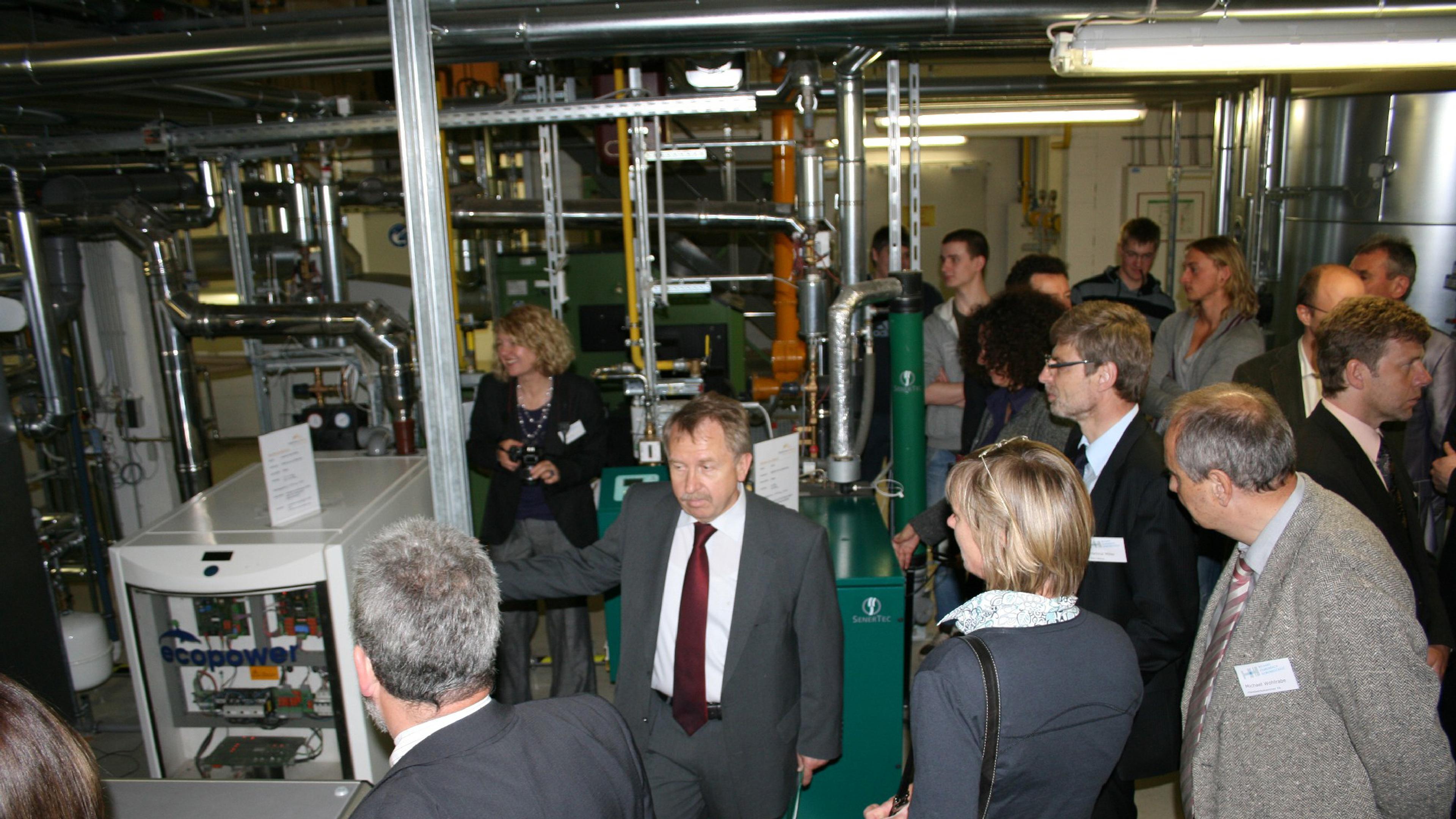Menschen in Anzügen und Geschäftskleidung versammeln sich um Industriemaschinen, um an einer Vorführung oder einer Führung in einer Technologieeinrichtung teilzunehmen.