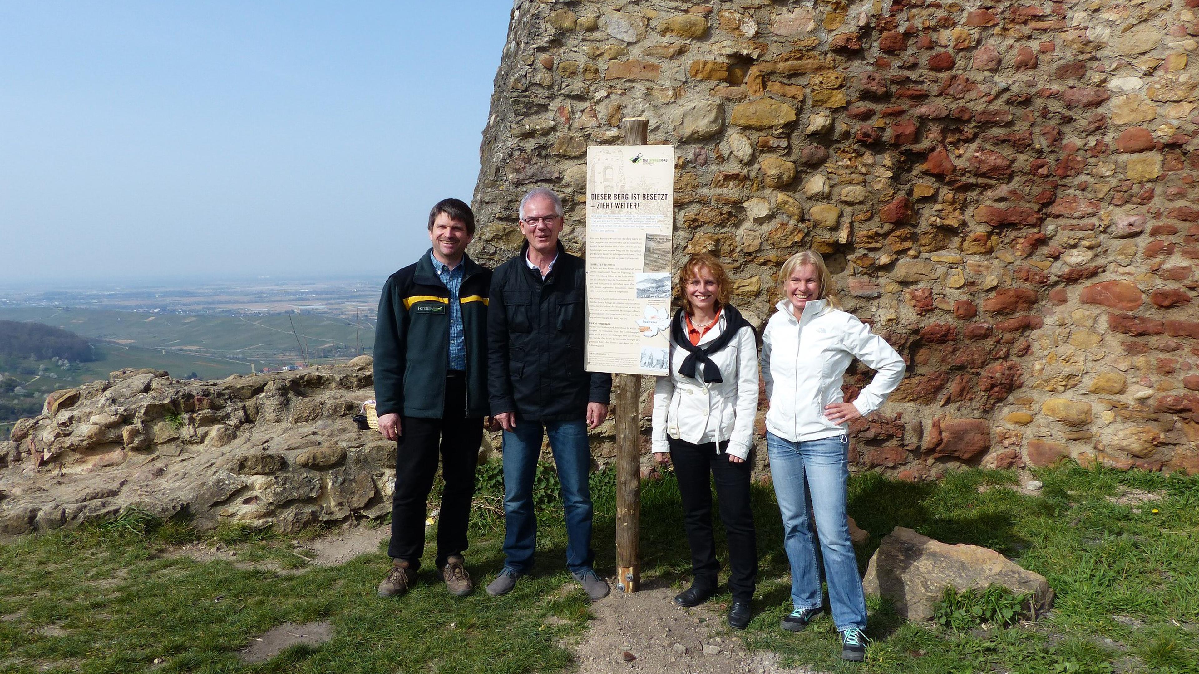 Vier Personen stehen vor einem Schild an einer Steinmauer, im Hintergrund bietet sich ein malerischer Blick auf sanfte Hügel.