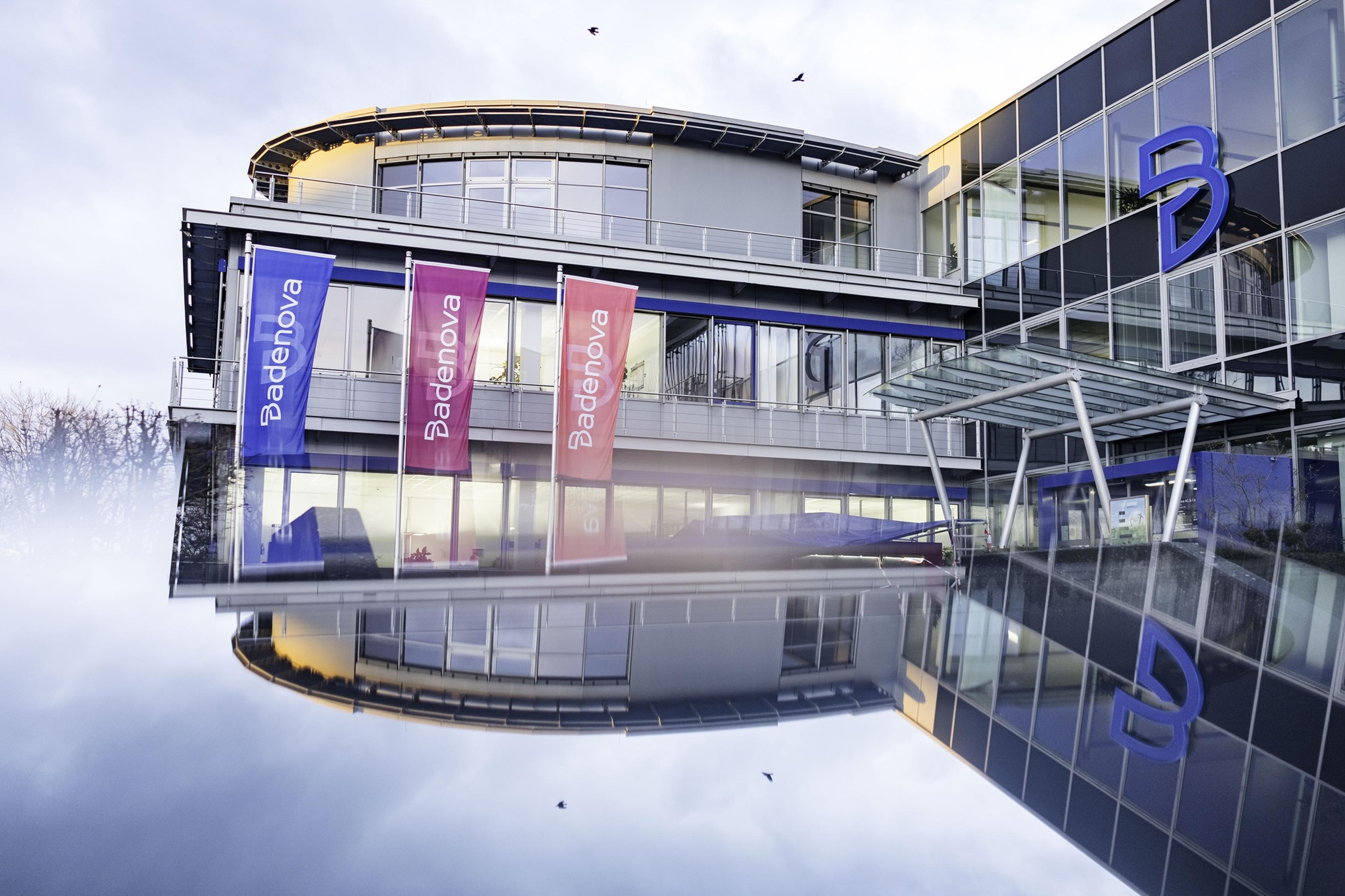Modern building with glass facade, colorful Badenova banners, and a large blue 'B' logo, reflected on a water surface.