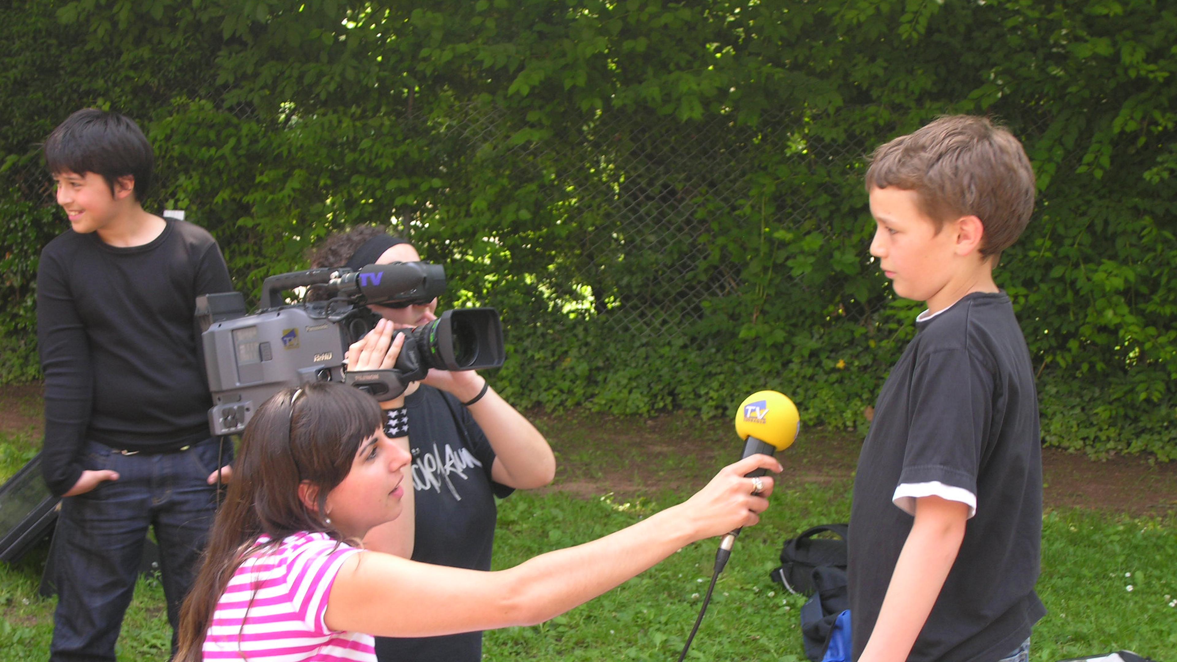Ein Junge in einem schwarzen Hemd wird von einer Frau mit einem Mikrofon interviewt, während eine andere Person draußen eine Videokamera bedient.