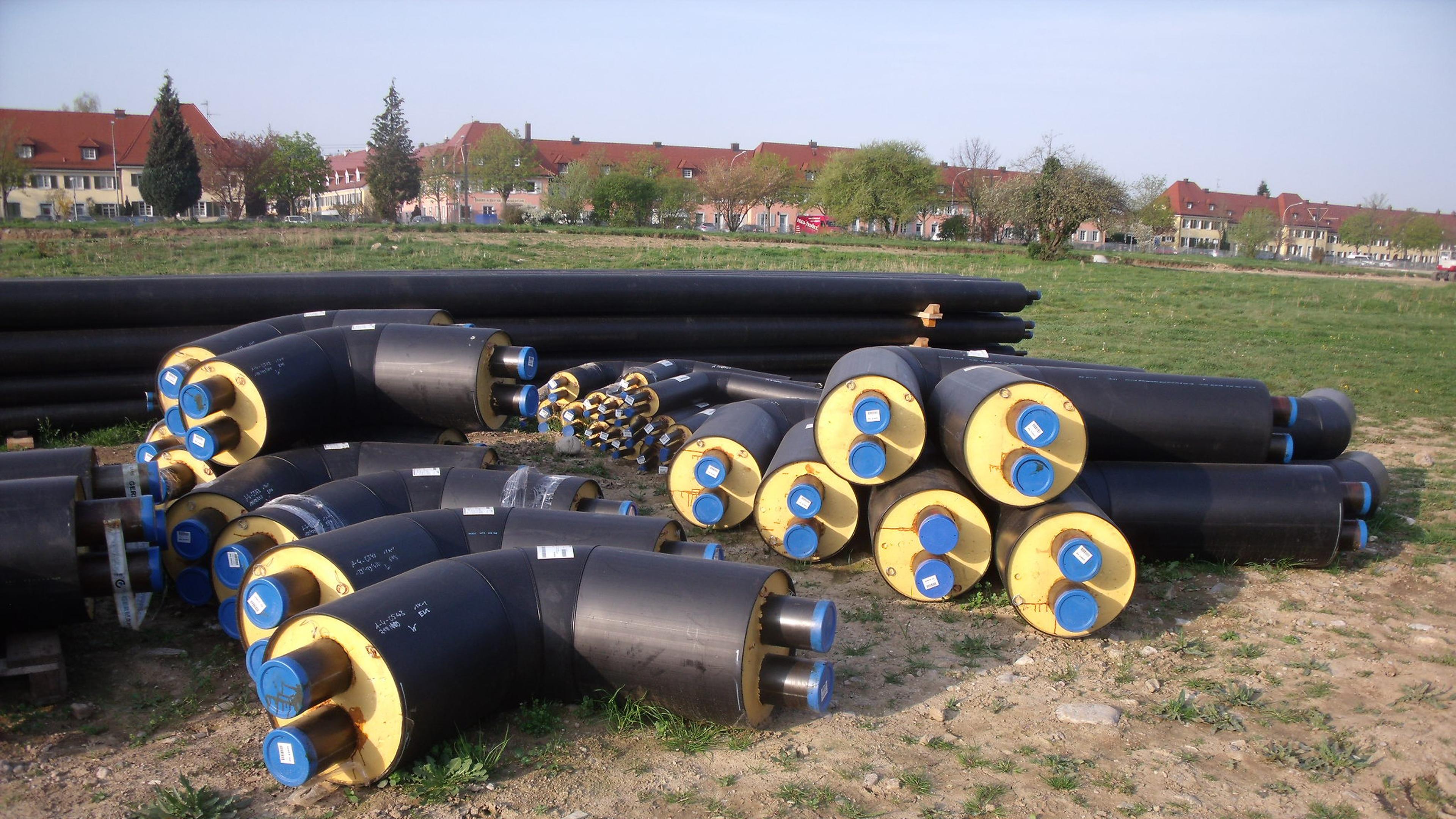 Stapel großer isolierter Rohre mit gelben und blauen Enden, die auf Gras und Erde liegen, im Hintergrund sind in der Ferne Häuser und Bäume zu sehen.