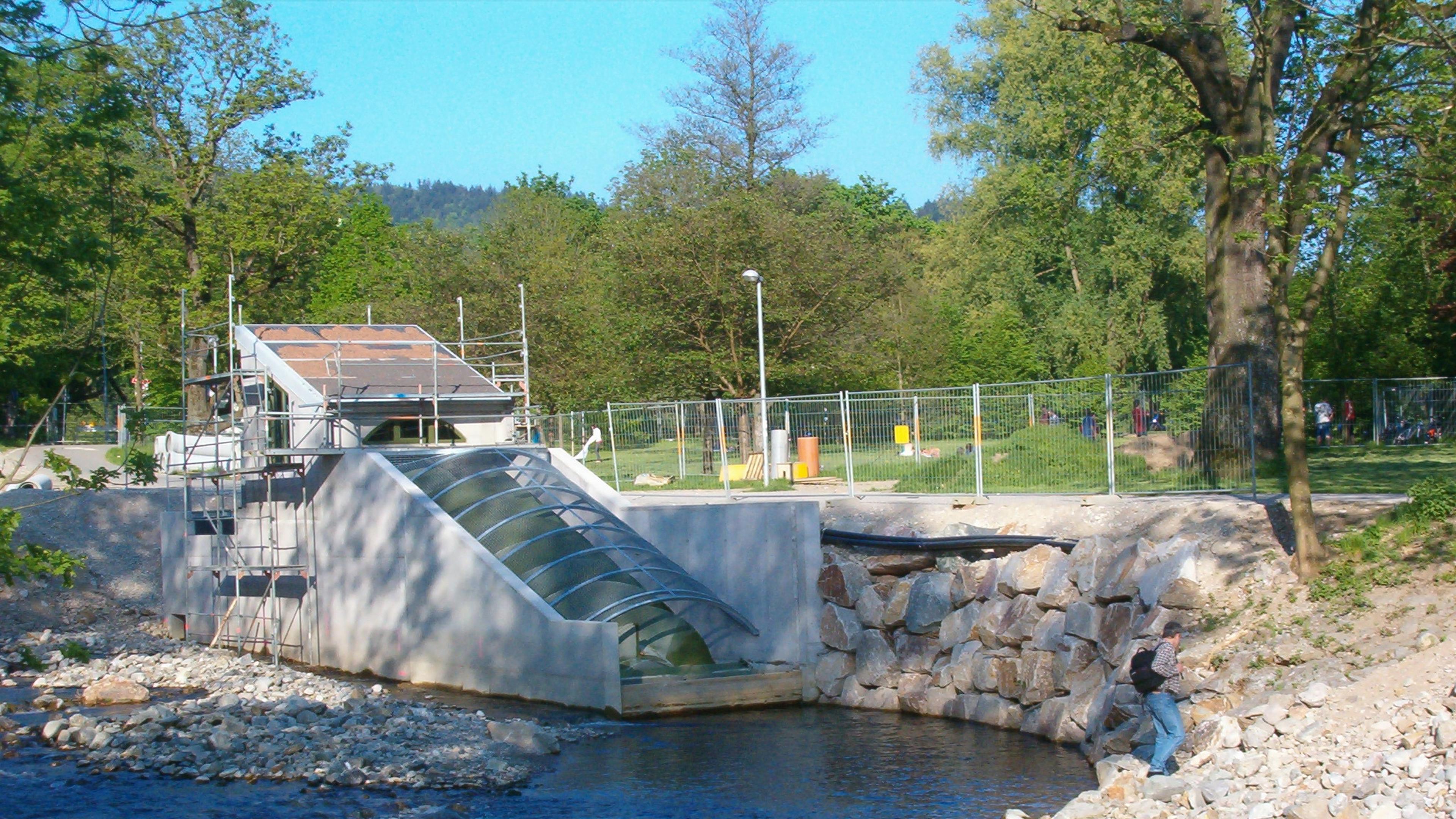 Ein kleines Wasserkraftwerk, das gerade an einem Fluss gebaut wird, umgeben von Bäumen und einem Zaun, mit einer Person, die in der Nähe spazieren geht.