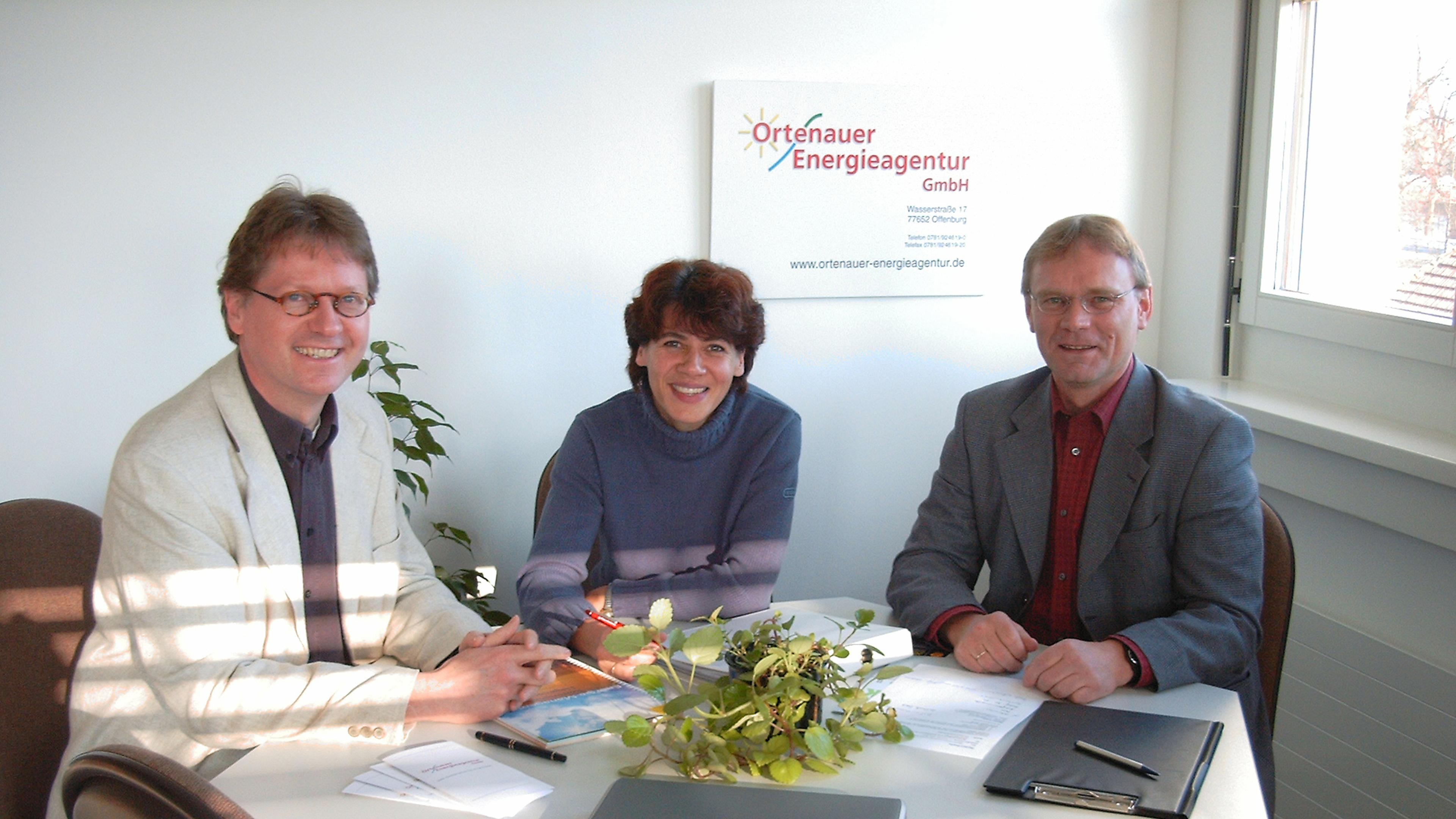 Drei Personen sitzen an einem Schreibtisch, umgeben von Unterlagen und einer Pflanze, und lächeln in die Kamera. An der Wand hängt ein Schild der Ortenauer Energieagentur GmbH.