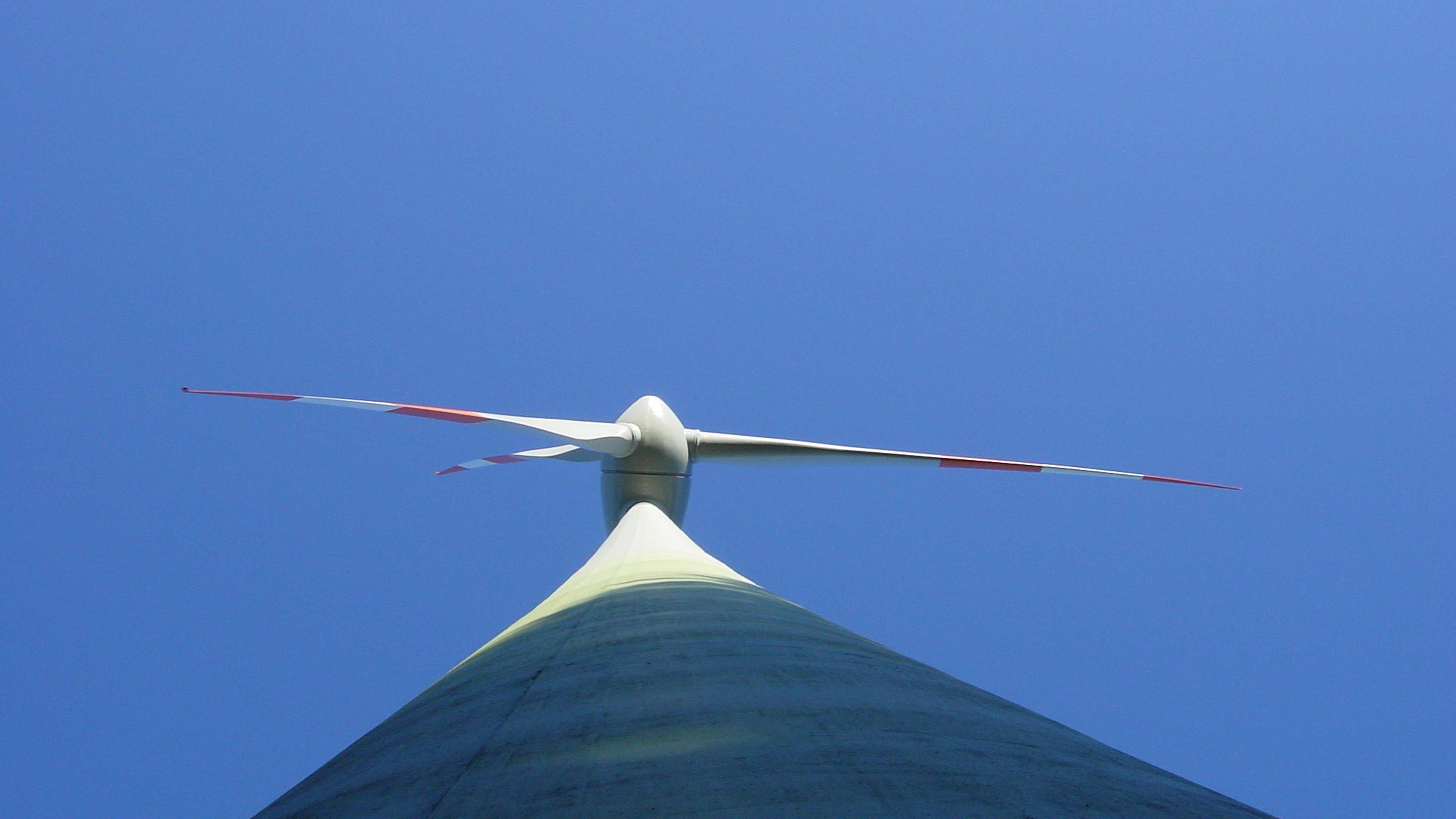 Eine Aufnahme aus niedriger Perspektive einer Windkraftanlage vor strahlend blauem Himmel, auf der zu sehen ist, wie sich ihre Rotorblätter horizontal vom Nabenbereich aus erstrecken.