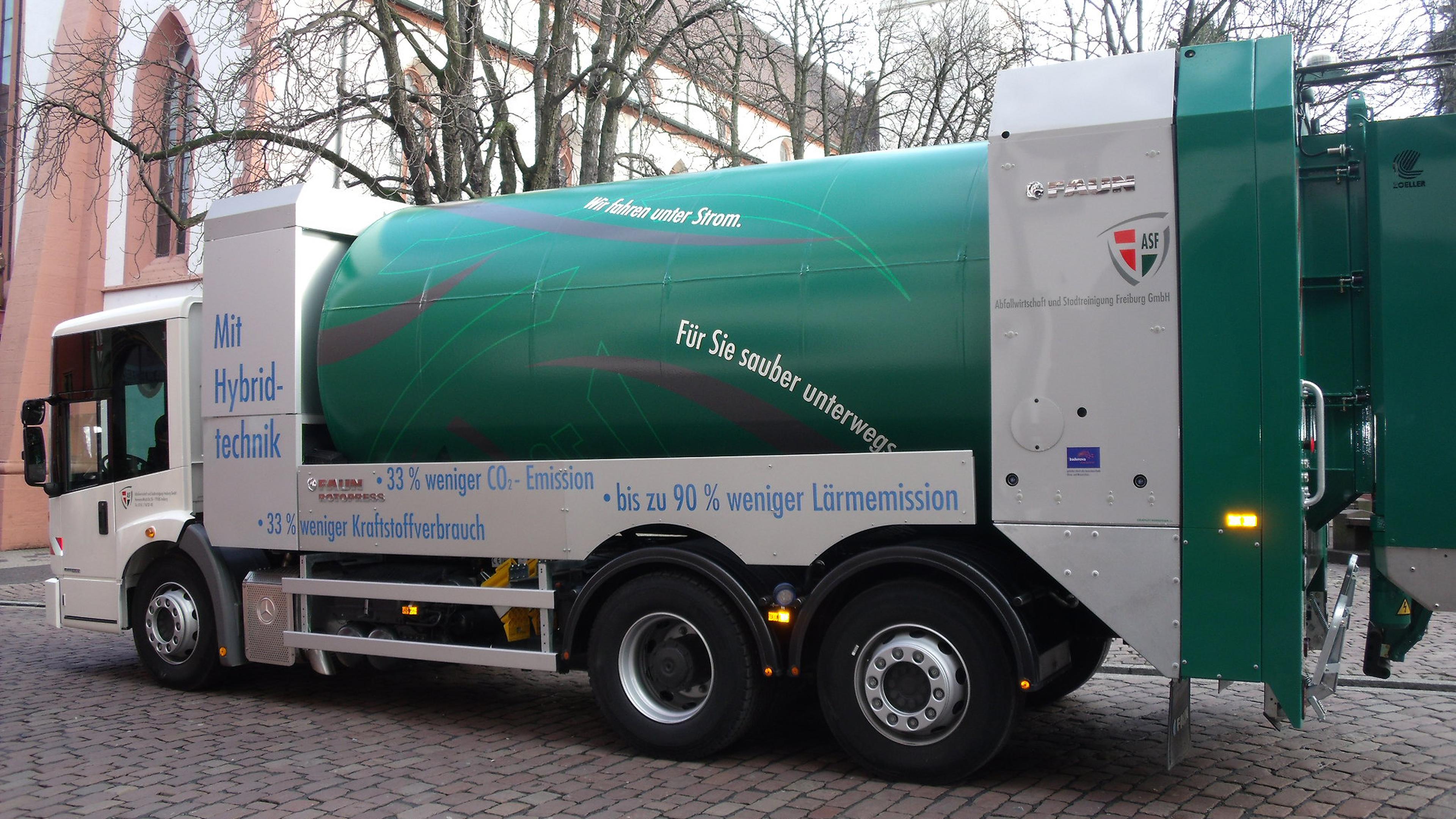 Ein grüner Hybrid-Müllwagen auf einer Kopfsteinpflasterstraße, versehen mit umweltfreundlichen Botschaften auf Deutsch zu reduzierten Emissionen und geringerem Kraftstoffverbrauch.