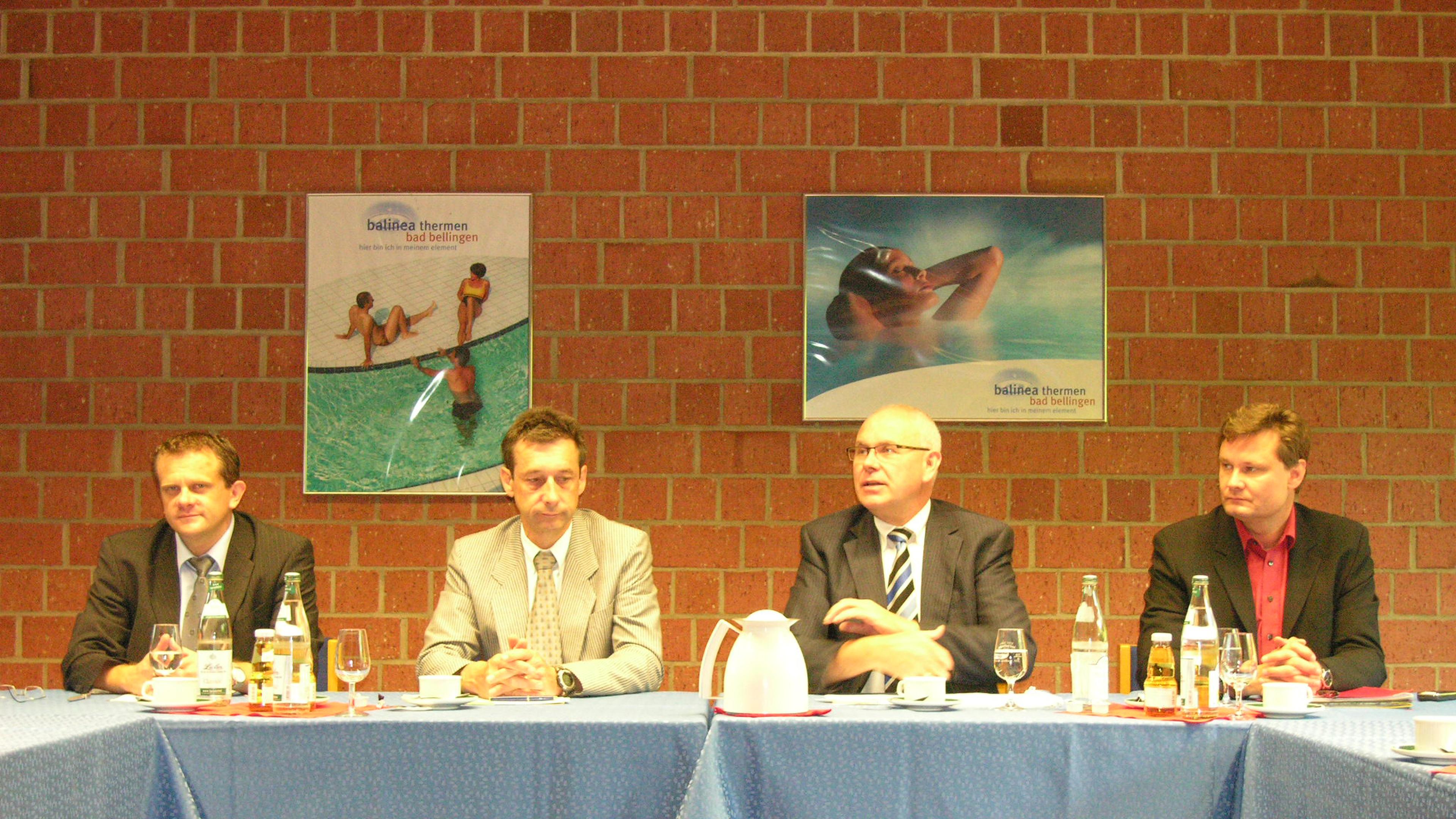 Vier Männer in Anzügen sitzen an einem Konferenztisch, auf dem Wasserflaschen und Gläser stehen, vor einer Backsteinwand mit Plakaten.