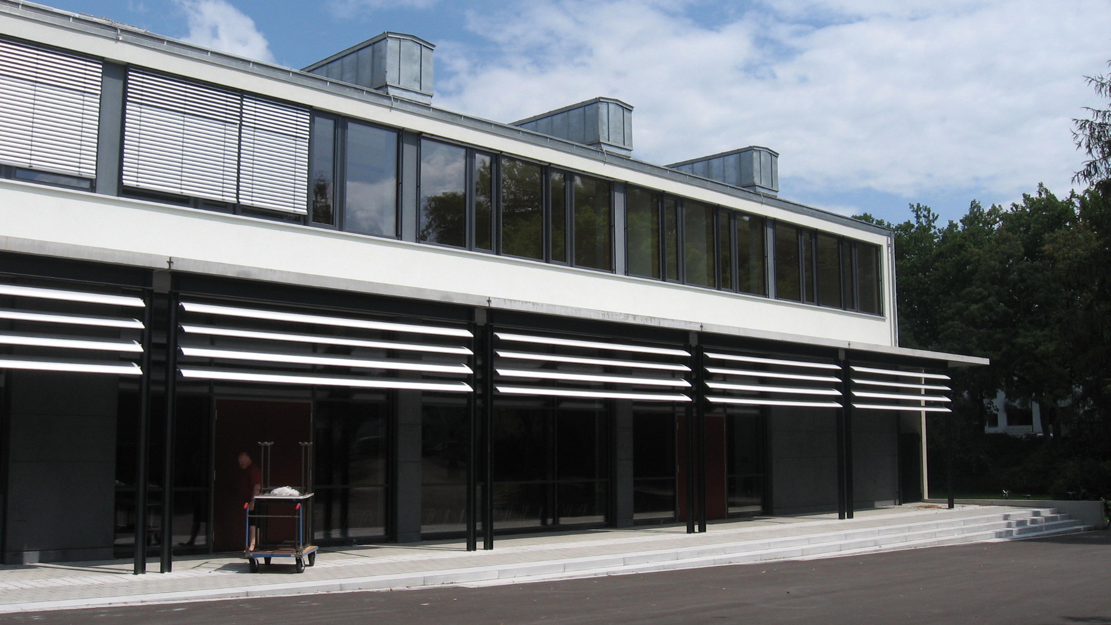 Modernes Gebäude mit großen Fenstern und horizontalen Außenlamellen, vor dem ein Wagen steht.