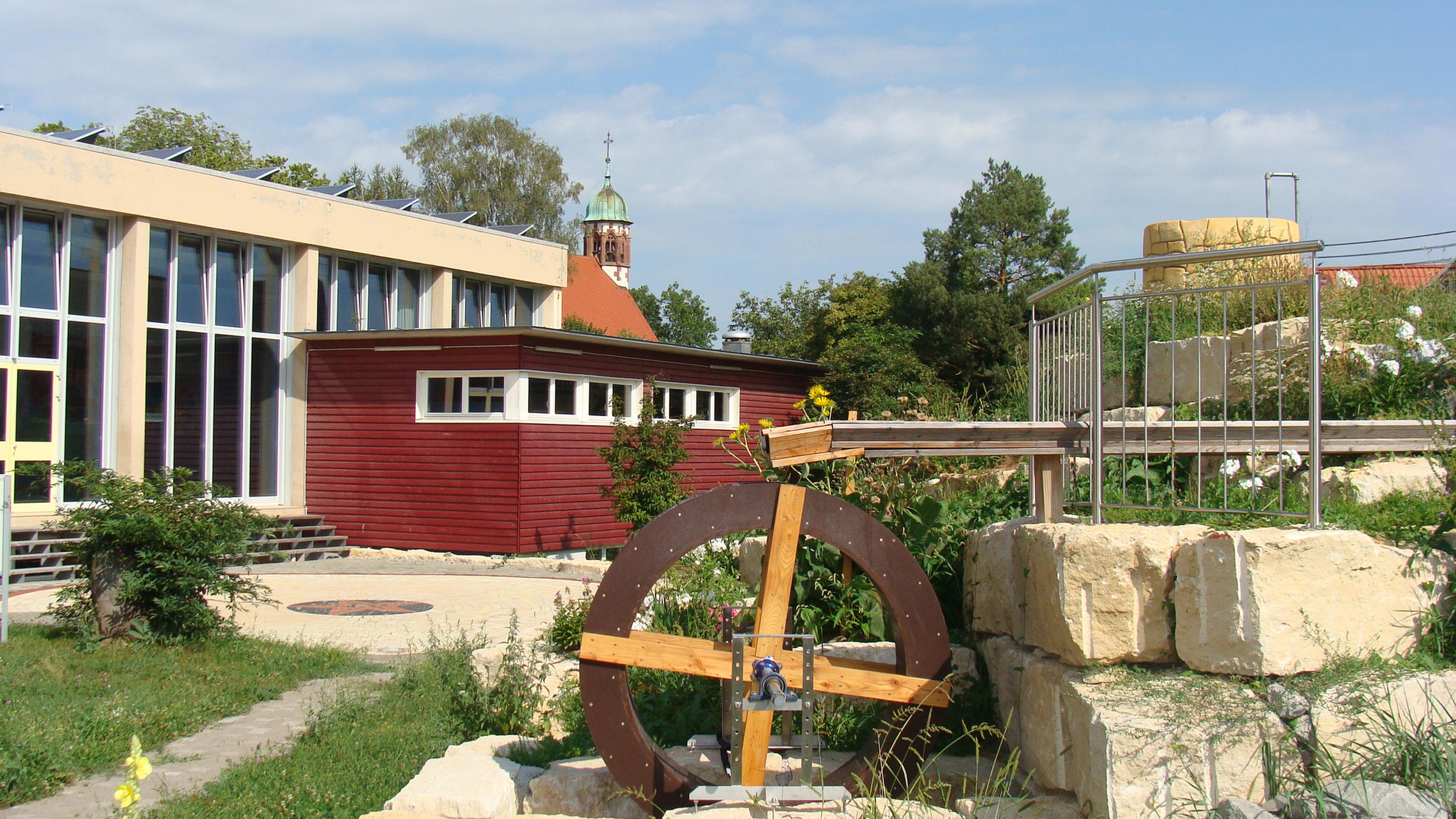 Ein modernes Gebäude mit großen Fenstern, einem roten Anbau und einem Wasserrad, umgeben von viel Grün. Im Hintergrund ist ein Turm zu sehen.