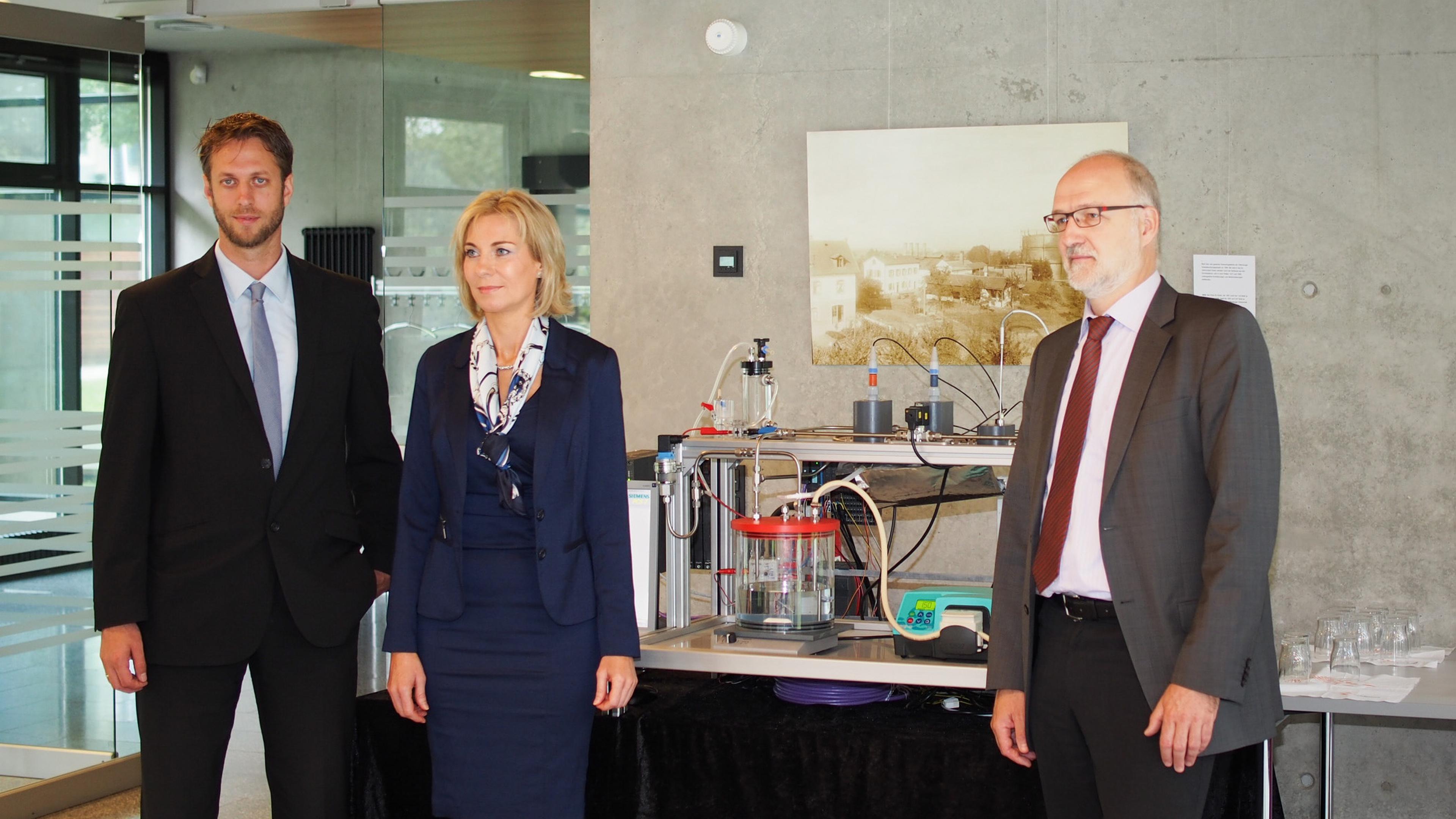 Three people in formal attire stand beside scientific equipment in a modern office setting.