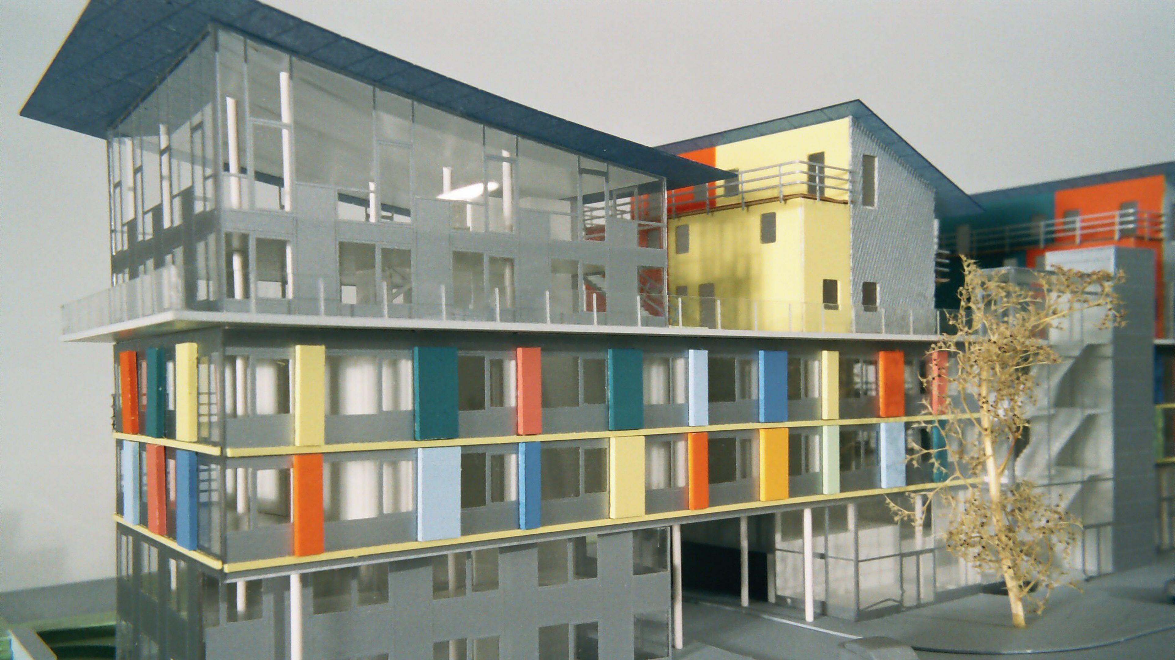 Architekturmodell eines modernen mehrstöckigen Gebäudes mit bunten Paneelen und großen Glasfenstern, vor dem ein kleiner Baum steht.