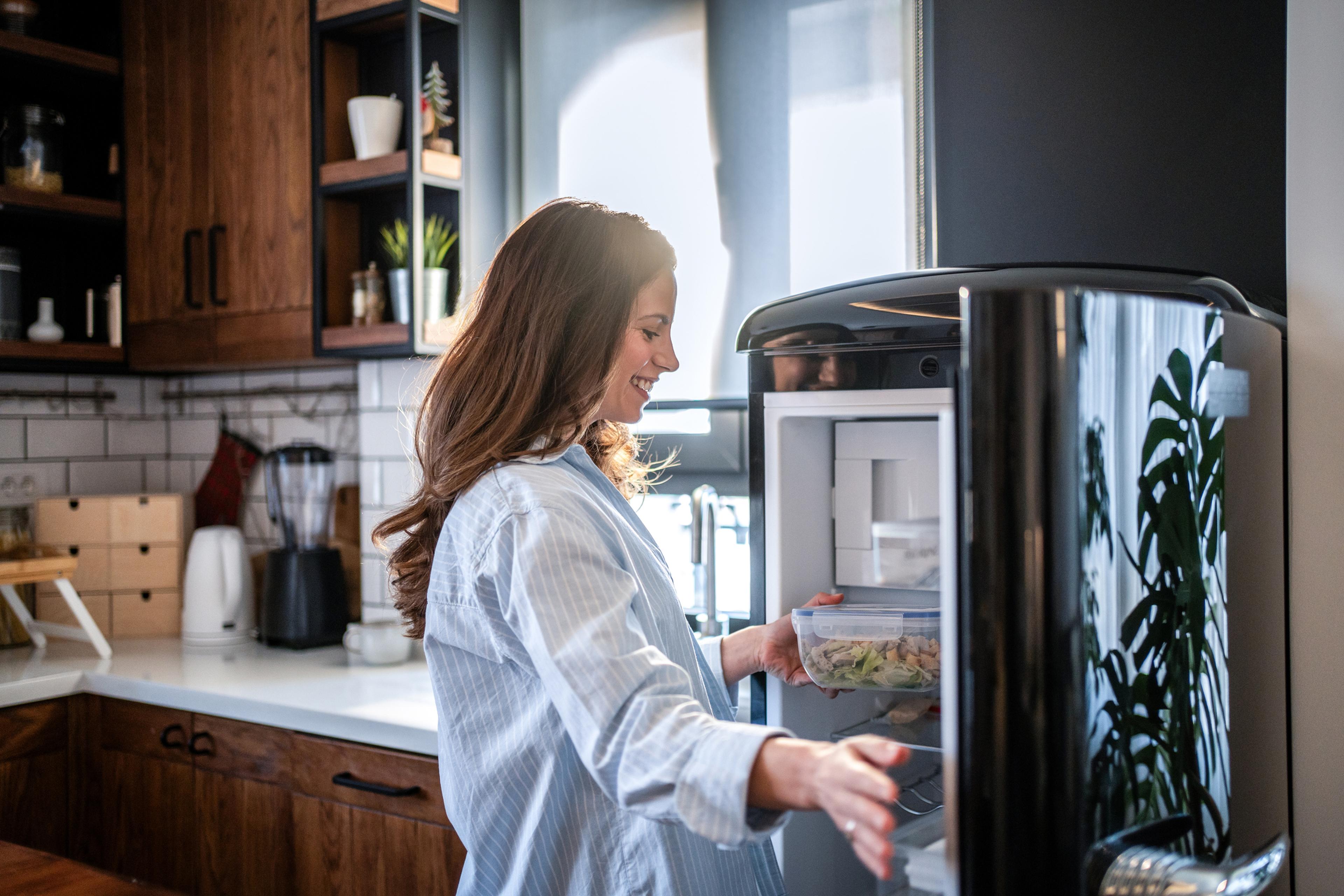 Eine Frau in einer gemütlichen Küche öffnet den Kühlschrank und lächelt, während sie einen Behälter mit Essen hineinstellt. Die Küche ist mit Holzschränken ausgestattet und modern eingerichtet.