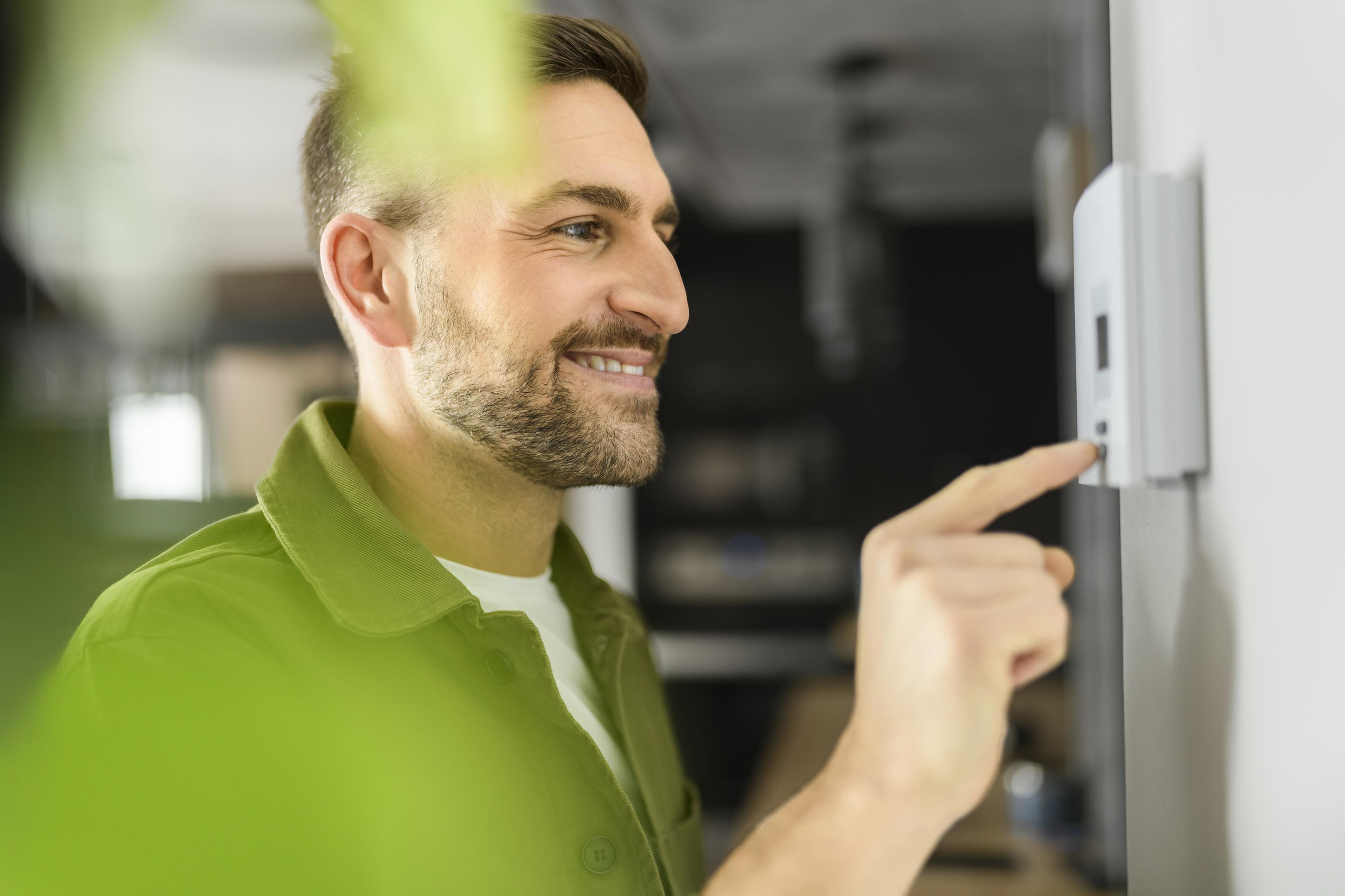 Ein Mann, der in einem modernen Wohnambiente an einem Thermostat an der Wand hantiert und dabei lächelt. Im Vordergrund ist eine grüne Pflanze zu sehen.