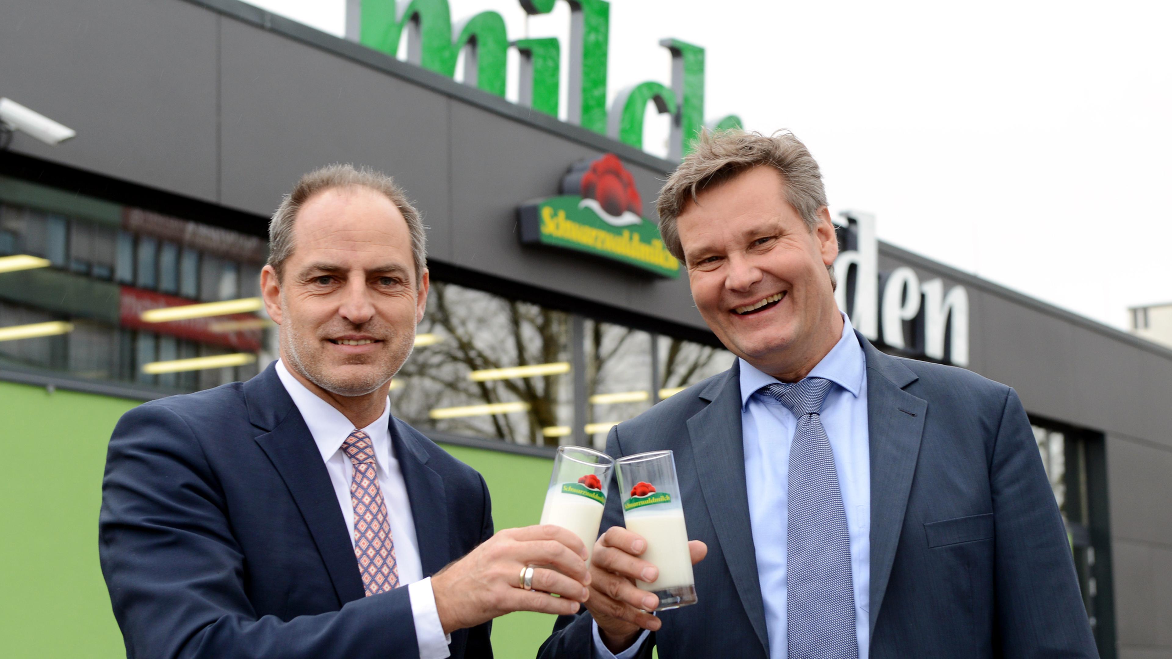 Zwei Männer in Anzügen stoßen mit Gläsern Milch vor einem Gebäude mit der Aufschrift „Milch“ an.