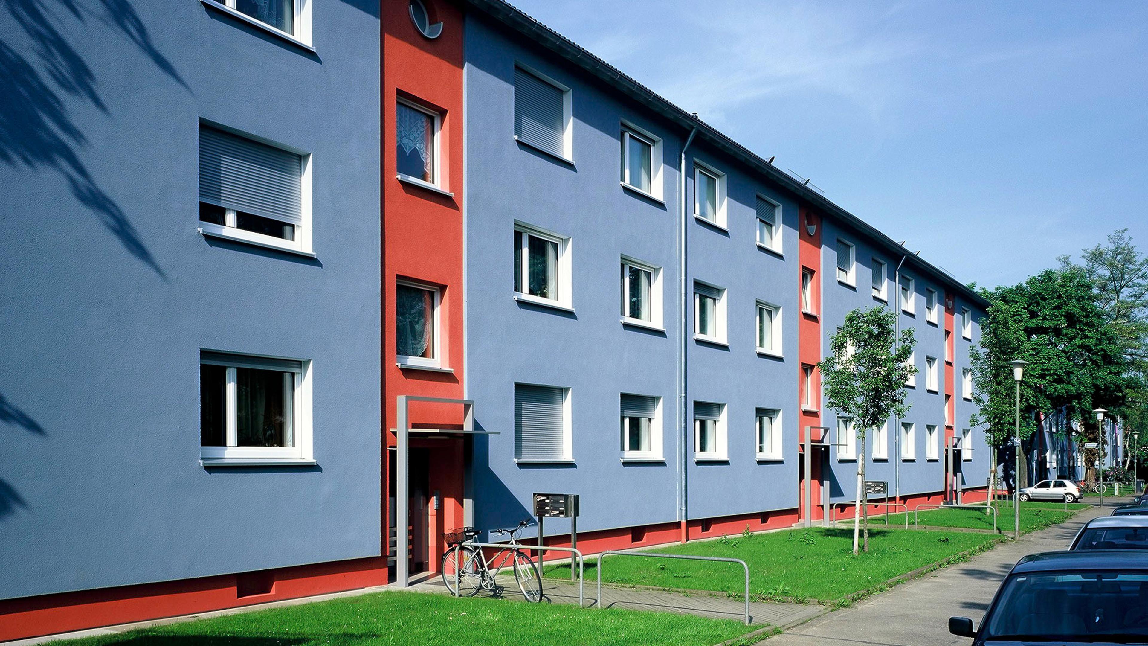 Blue and red apartment building with white-framed windows, bicycle parked outside, and a grassy lawn with small trees.