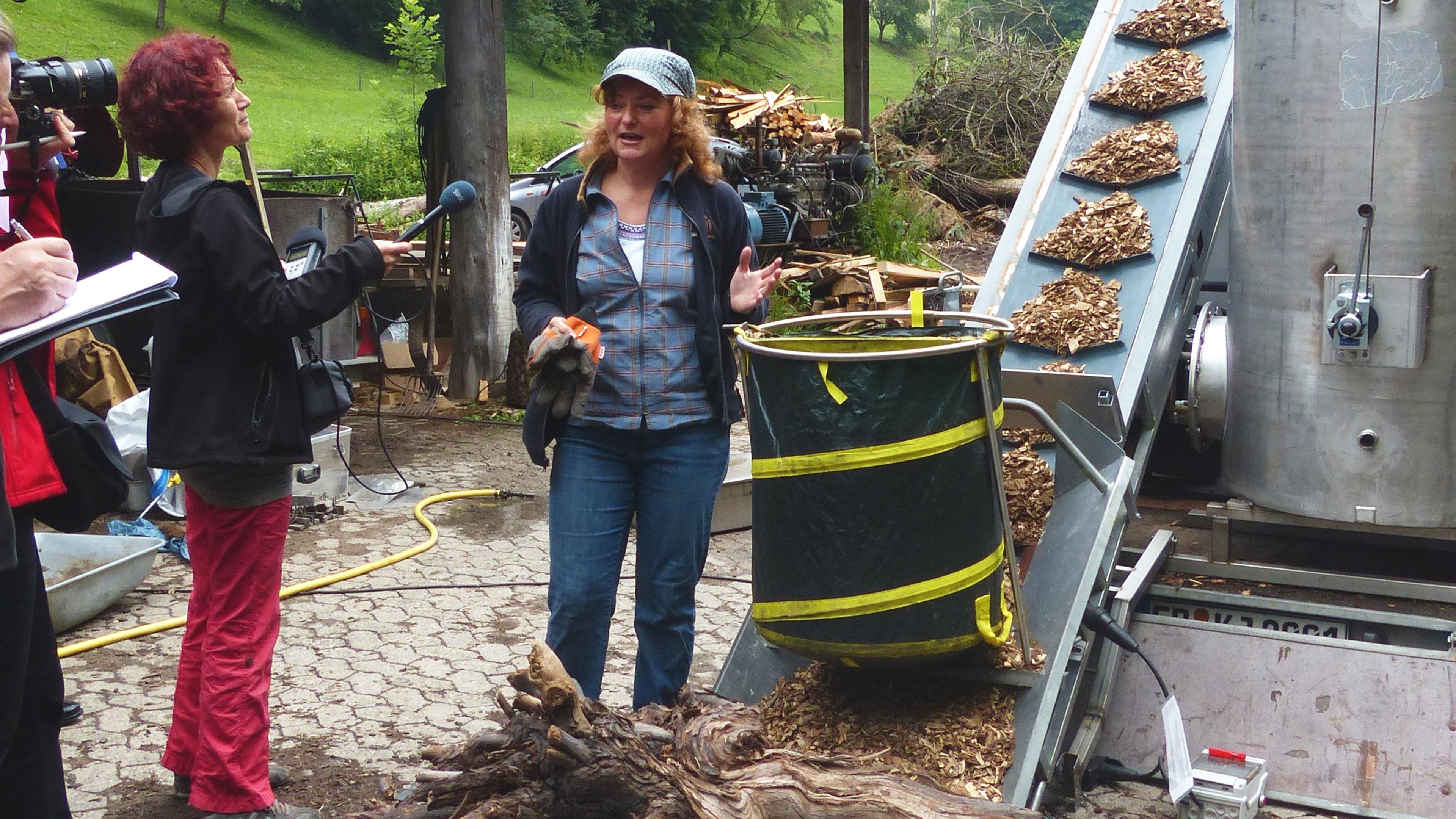 Eine Frau spricht mit Journalisten in der Nähe einer Holzverarbeitungsmaschine auf einem Bauernhof, während auf einem Förderband Holzspäne transportiert werden.