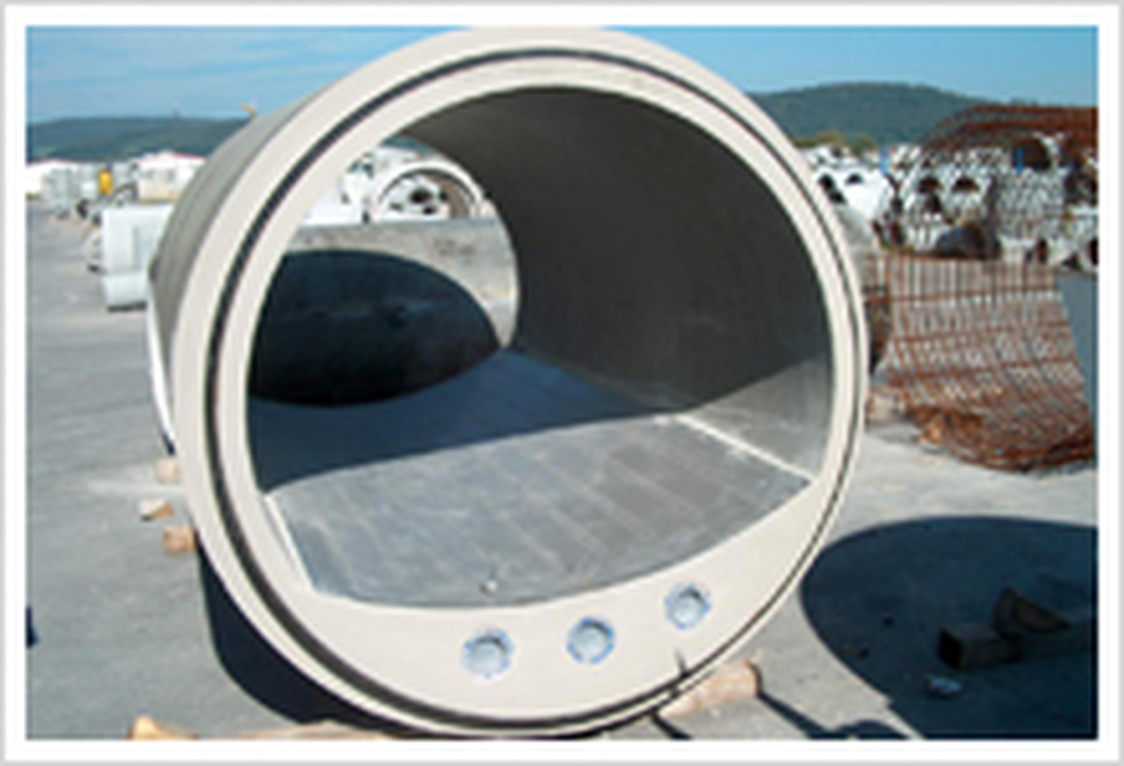 Ein großes Betonrohrsegment auf einer Baustelle, mit glatter Innenseite und drei kleinen Öffnungen unten vorne. Im Hintergrund sind Hügel zu sehen.