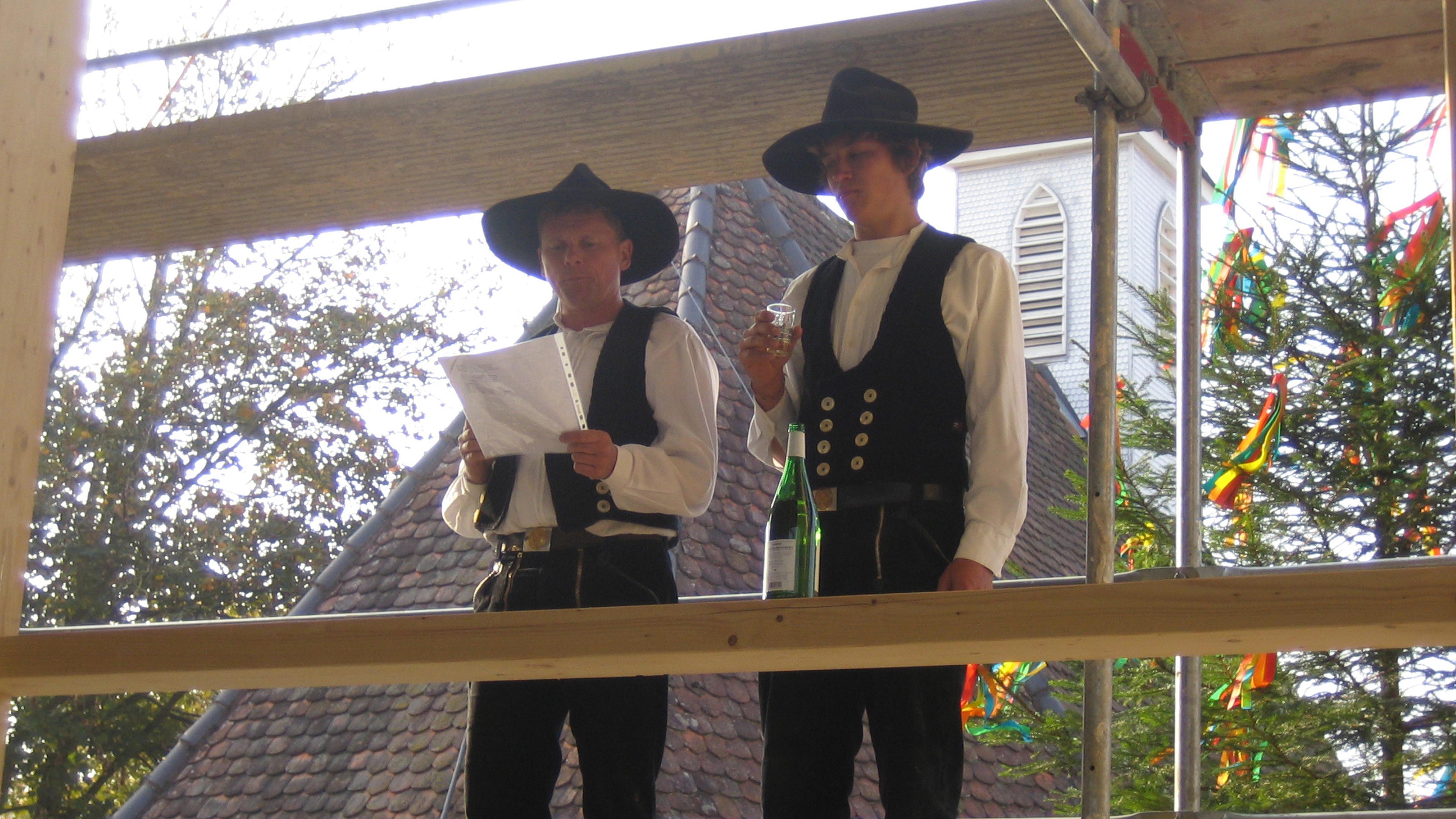 Zwei Maenner in traditioneller Tracht stehen auf einem Gerüst, der eine hält ein Blatt Papier in der Hand, der andere ein Glas; im Hintergrund sind ein festlich geschmückter Baum und ein Kirchturm zu sehen.