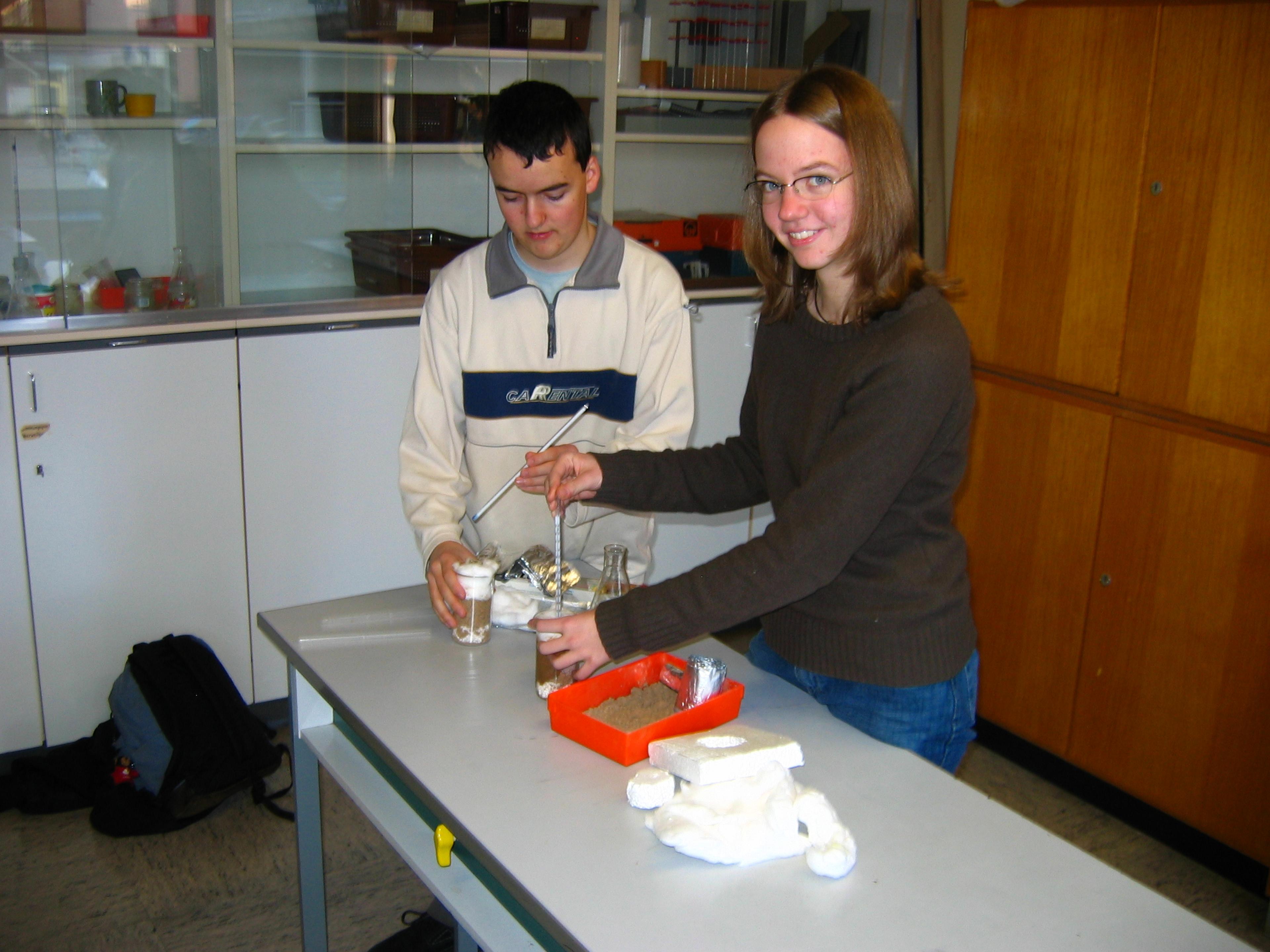 Zwei Personen führen an einem Tisch in einem Klassenzimmer ein wissenschaftliches Experiment durch und verwenden dabei verschiedene Materialien, darunter Bechergläser und Zangen.