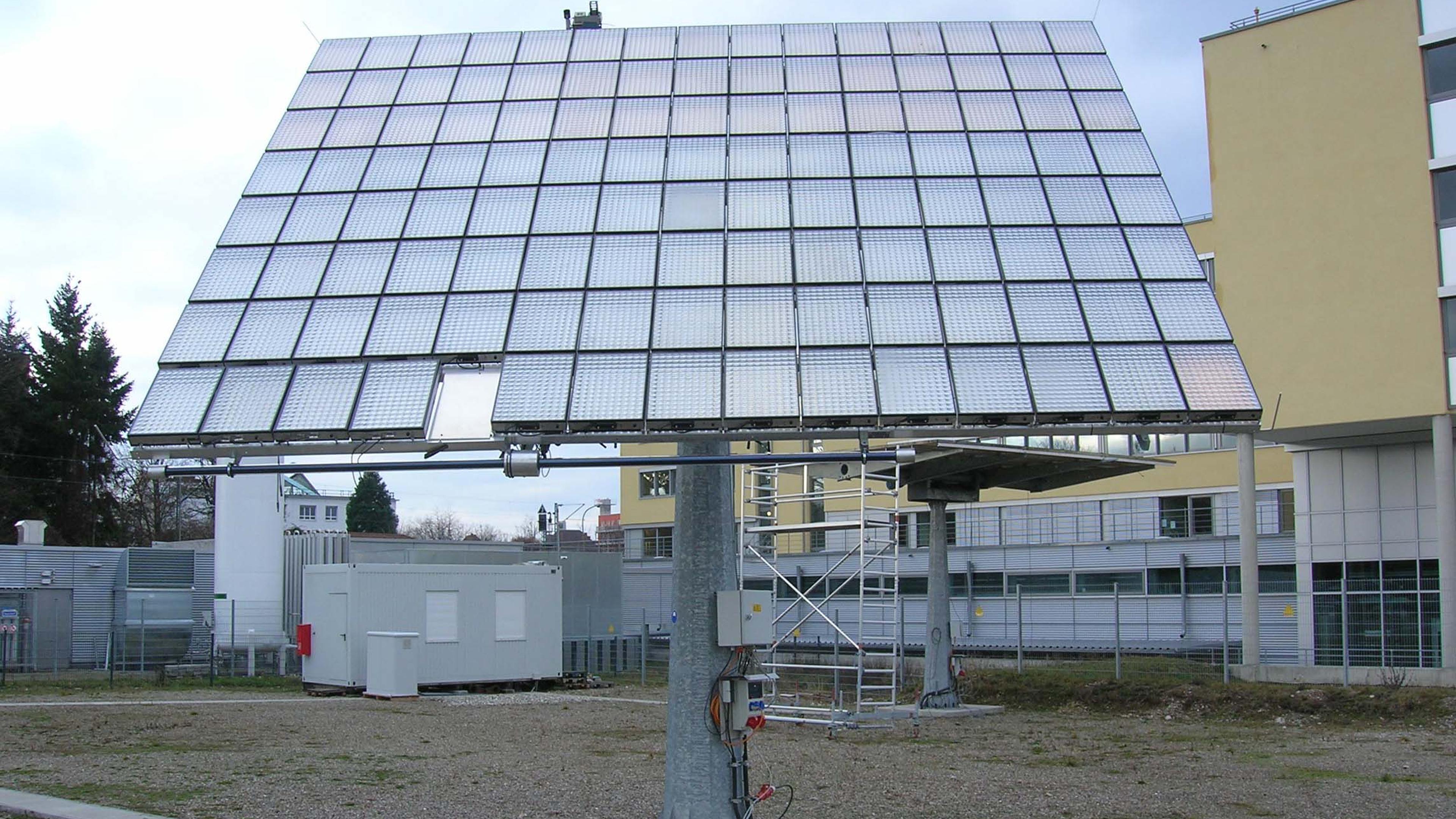 Eine große Solaranlage auf einem Metallgestell in einer Industriegegend, mit Gebäuden und Bäumen im Hintergrund unter einem bewölkten Himmel.