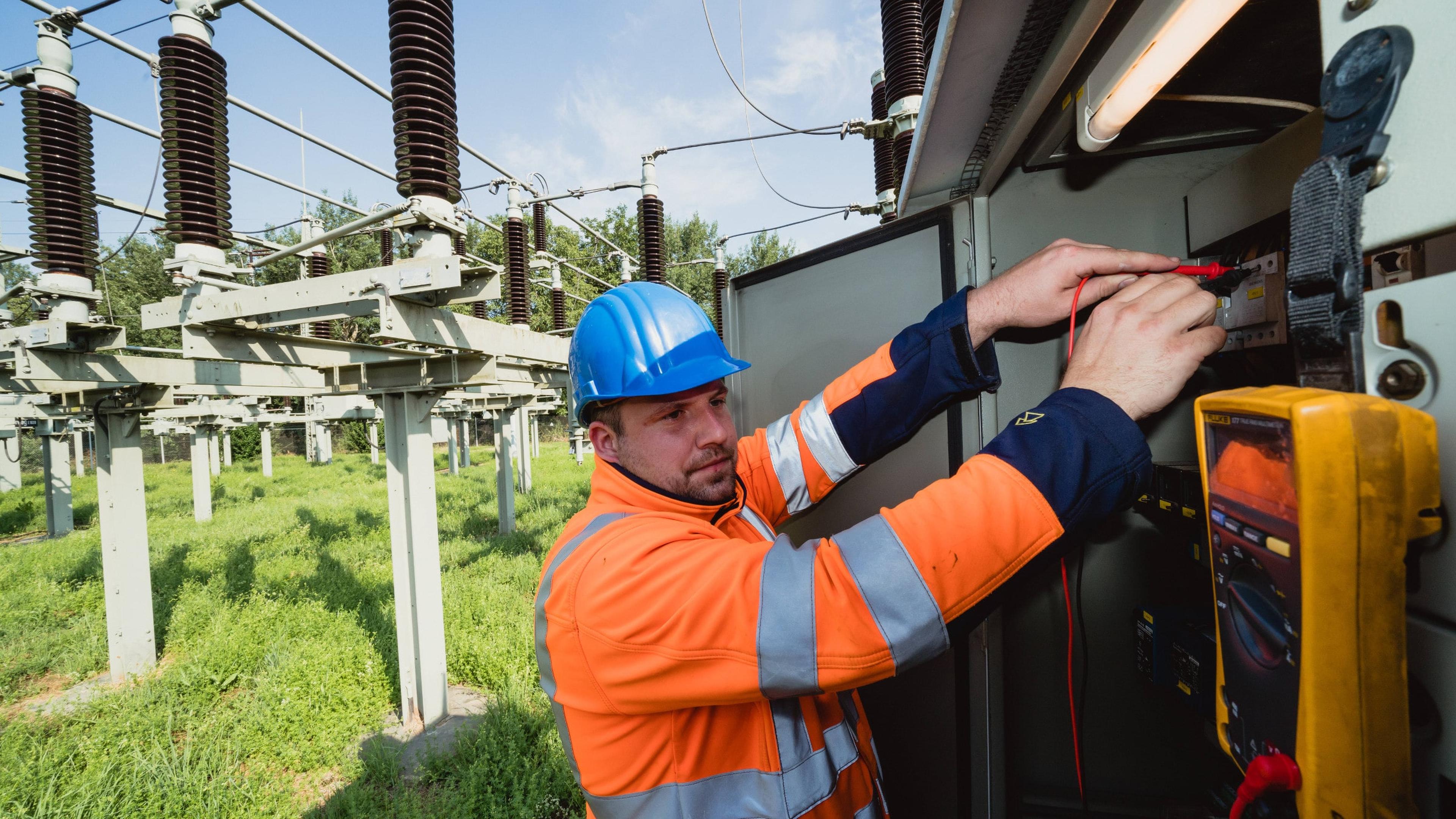 Techniker in orangefarbener Schutzkleidung und blauem Helm arbeitet an elektrischen Geräten in einem Freiluftkraftwerk.