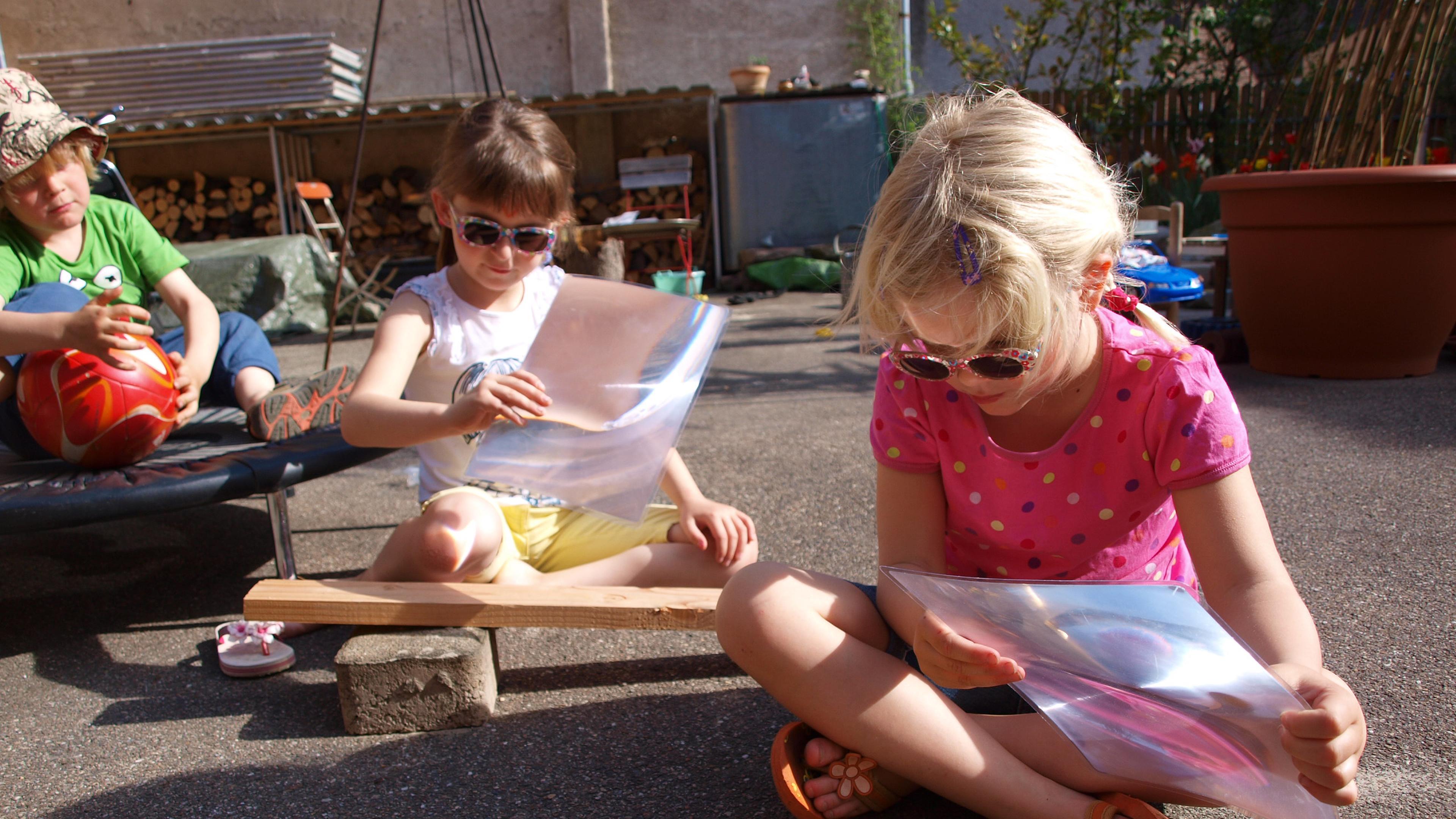 Kinder mit Sonnenbrillen benutzen an einem sonnigen Tag Lupe-Folien, waehrend sie auf dem Boden sitzen, im Hintergrund ein Sandkasten und Spielzeug.