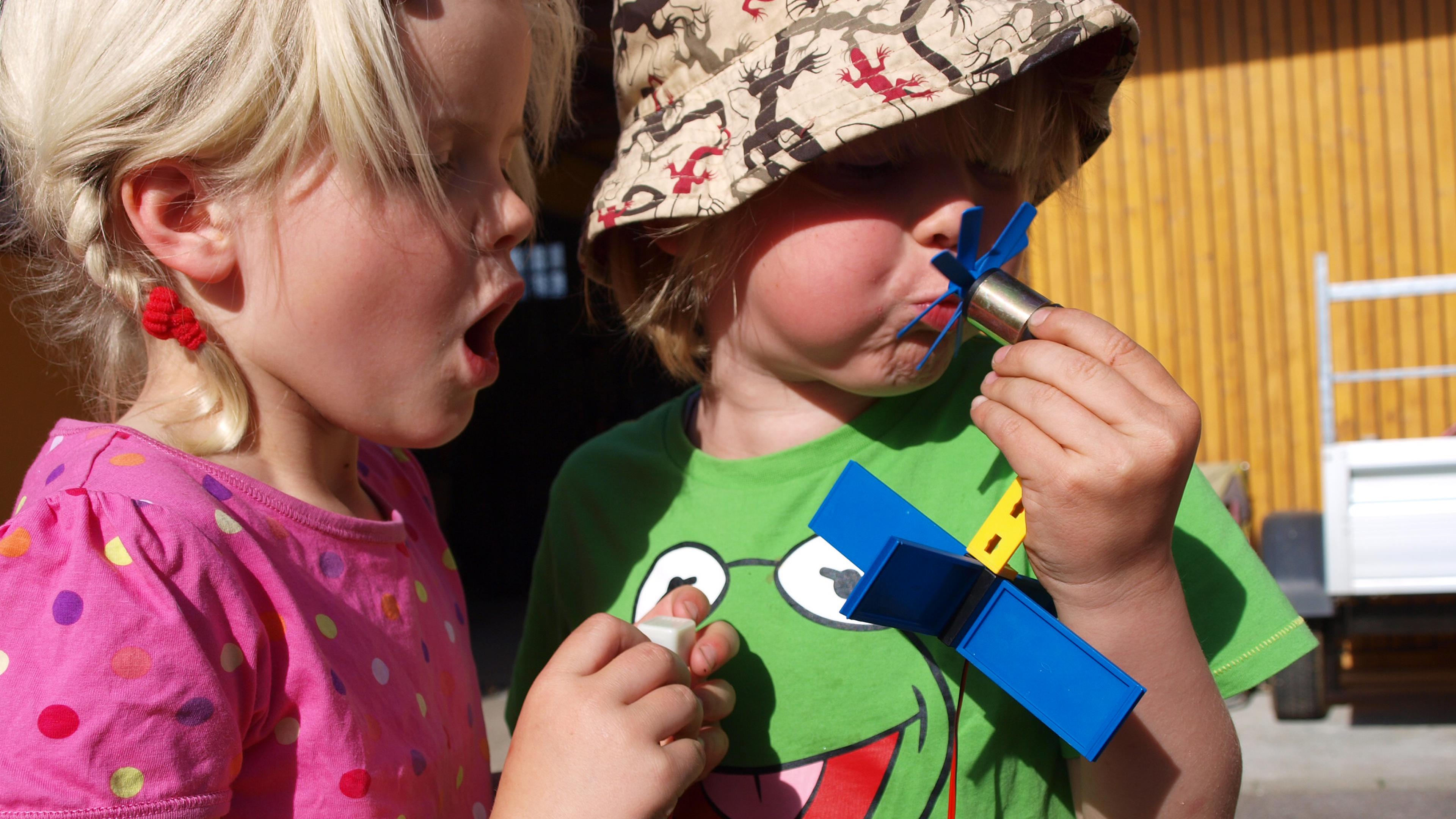 Zwei Kinder sind draußen. Das eine trägt einen Hut und spielt mit einem Spielzeug, während das andere in einem rosa gepunkteten Hemd überrascht zusieht.