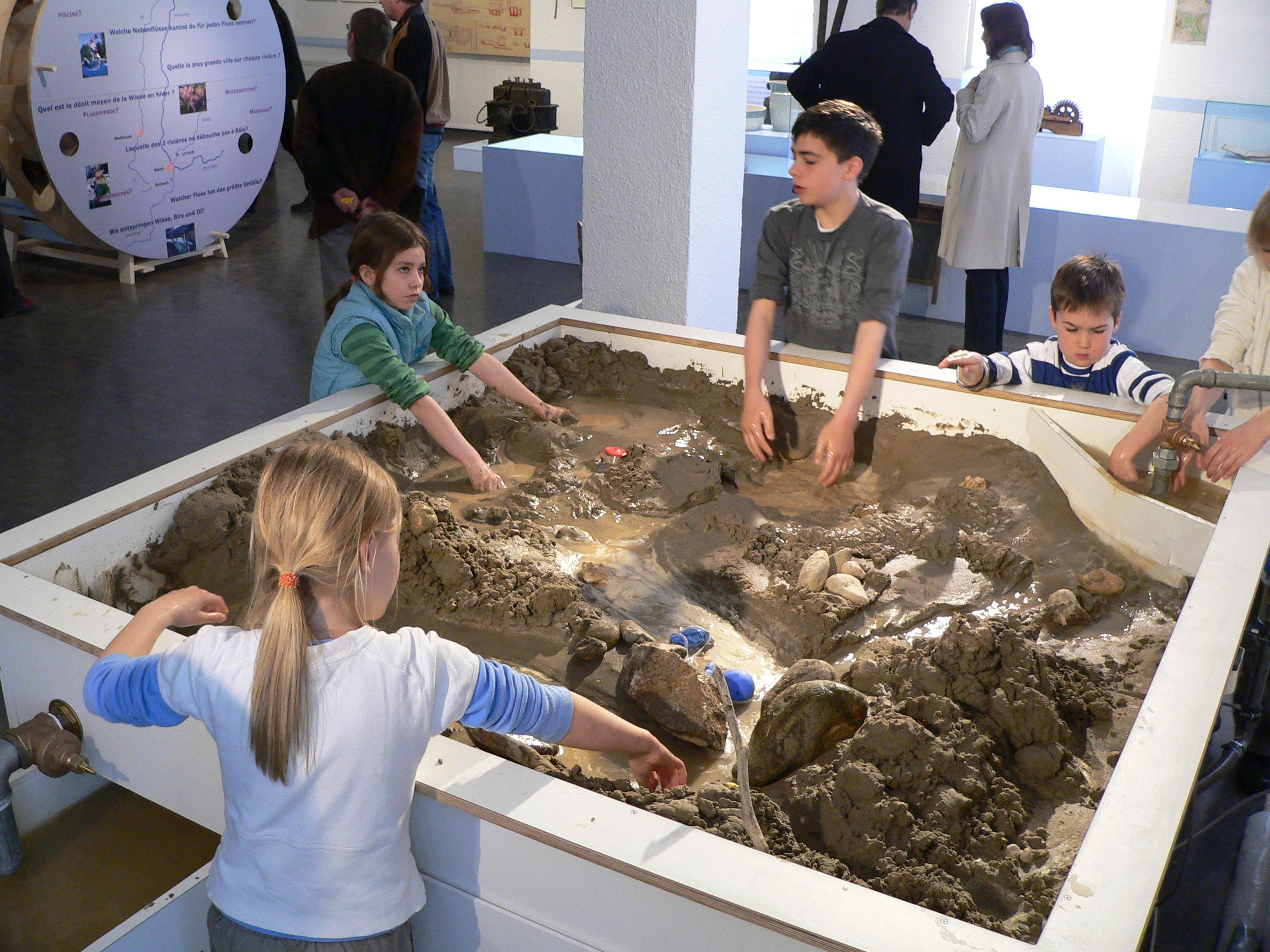 Kinder spielen in einer Indoor-Ausstellung mit Schlamm und Wasser und gestalten Landschaften in einem großen interaktiven Sandkasten.